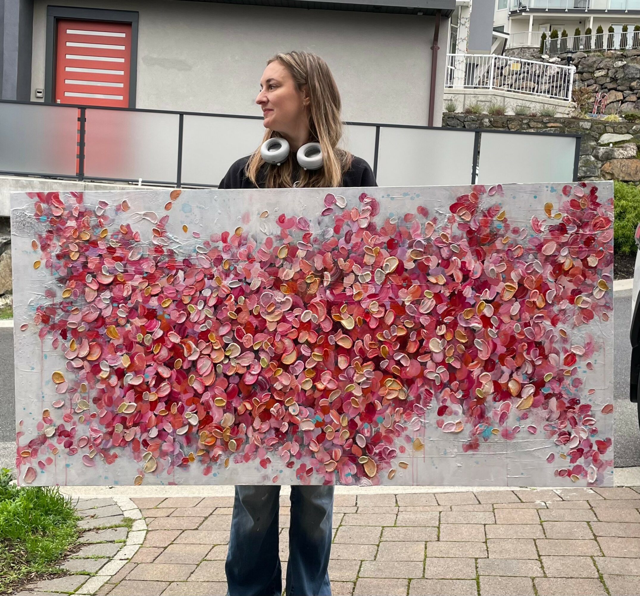 Woman holding a large colorful heart-shaped artwork outdoors, with a building and stone wall in background.
