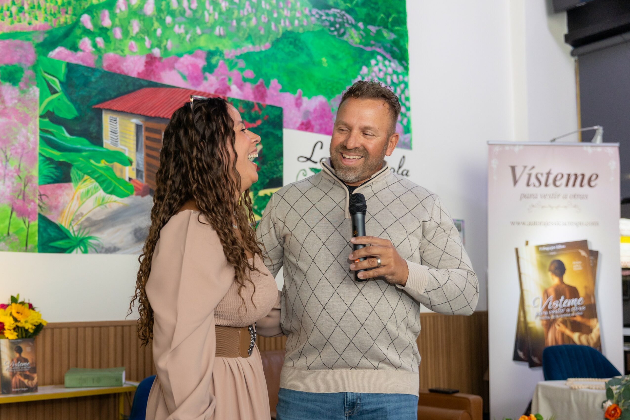 A man and woman are smiling and talking, with the man holding a microphone, in front of a colorful painting and a poster.