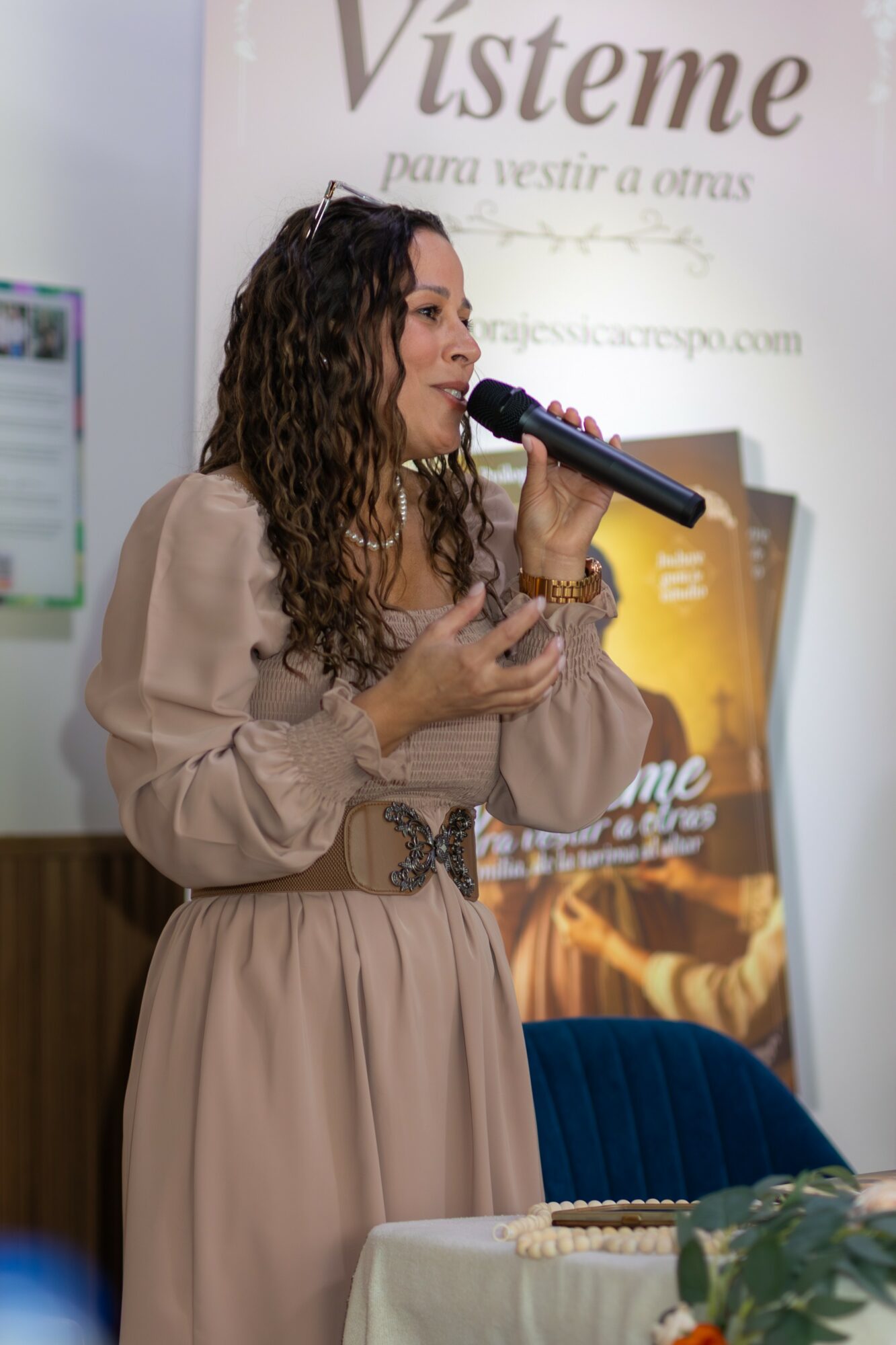 Woman with curly hair speaking into a microphone at an event, standing near a table with a plant and papers.