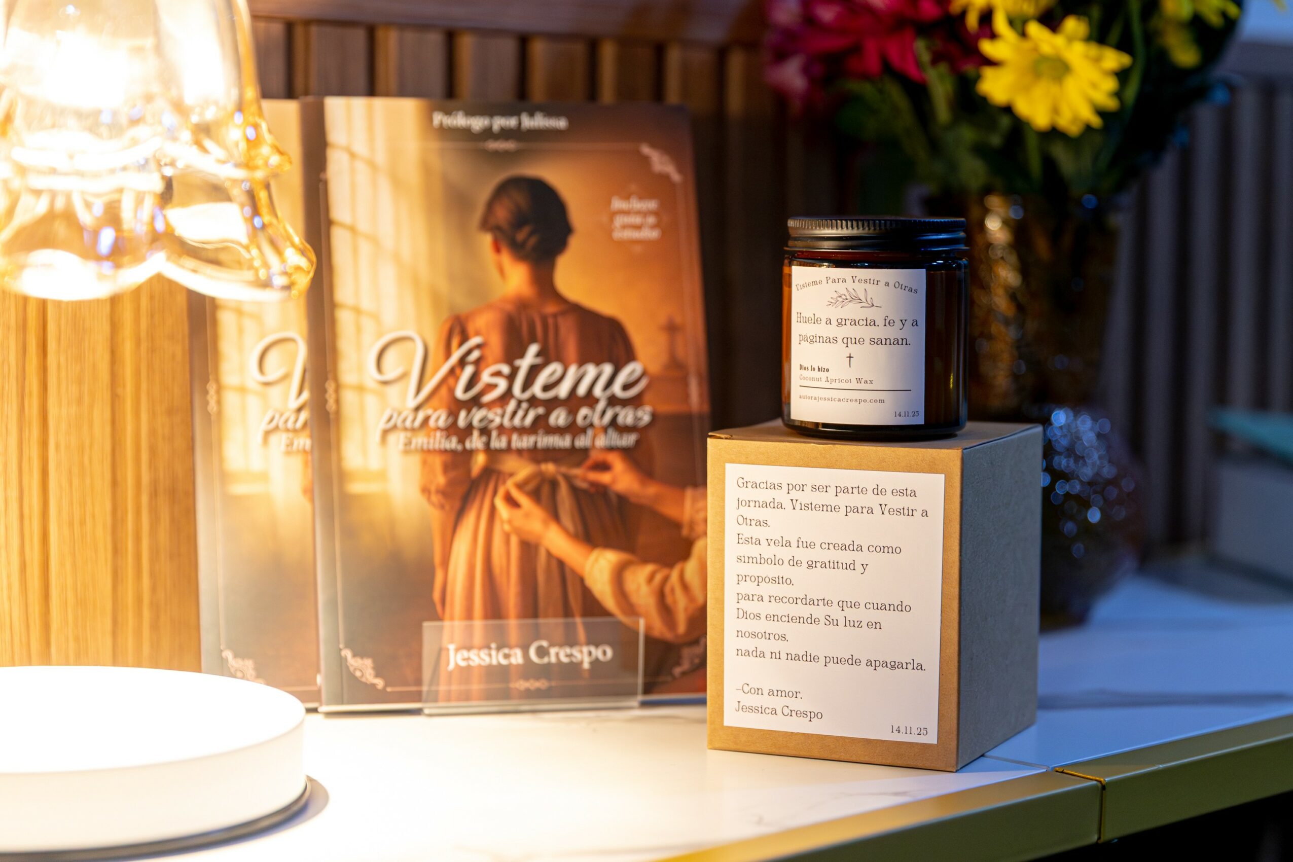 Open book titled 'Vísteme' with a woman on the cover, next to a candle, a jar, and a box on a table, with flowers in the background.