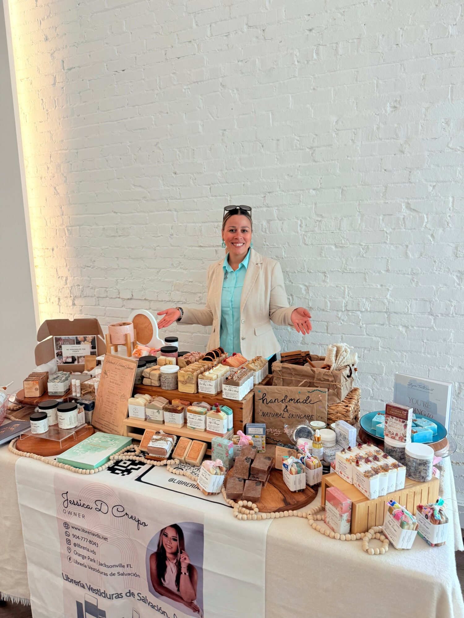 Person standing behind a table with various soap and skincare products, smiling, against a plain wall.