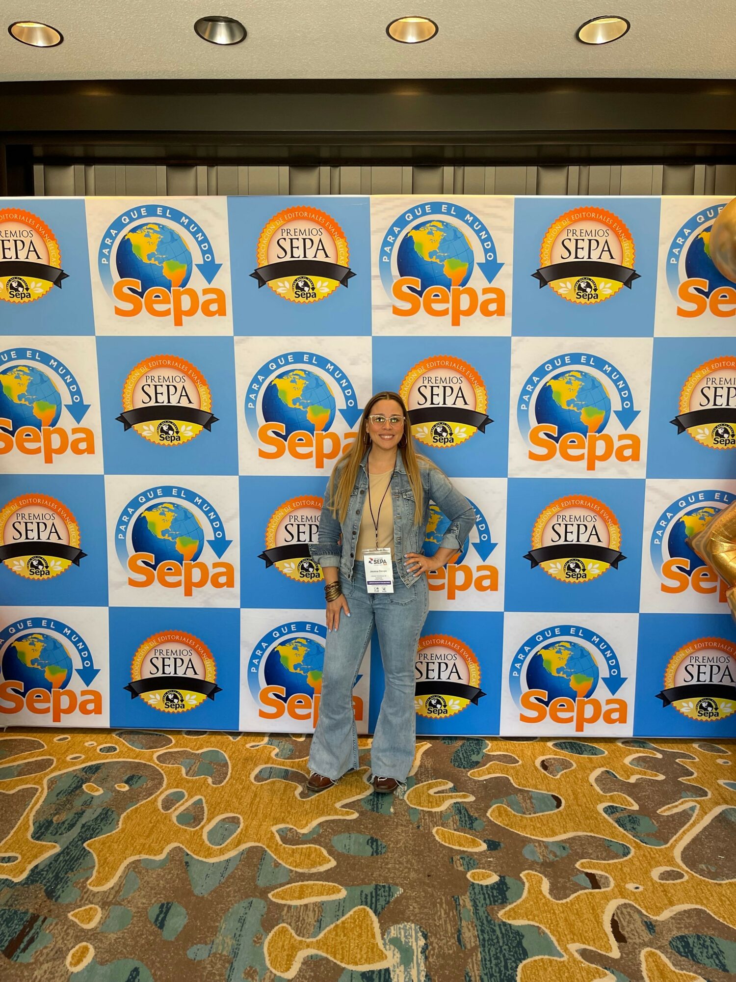 Person standing in front of a blue and orange SePa backdrop with logos, on a patterned carpeted floor.