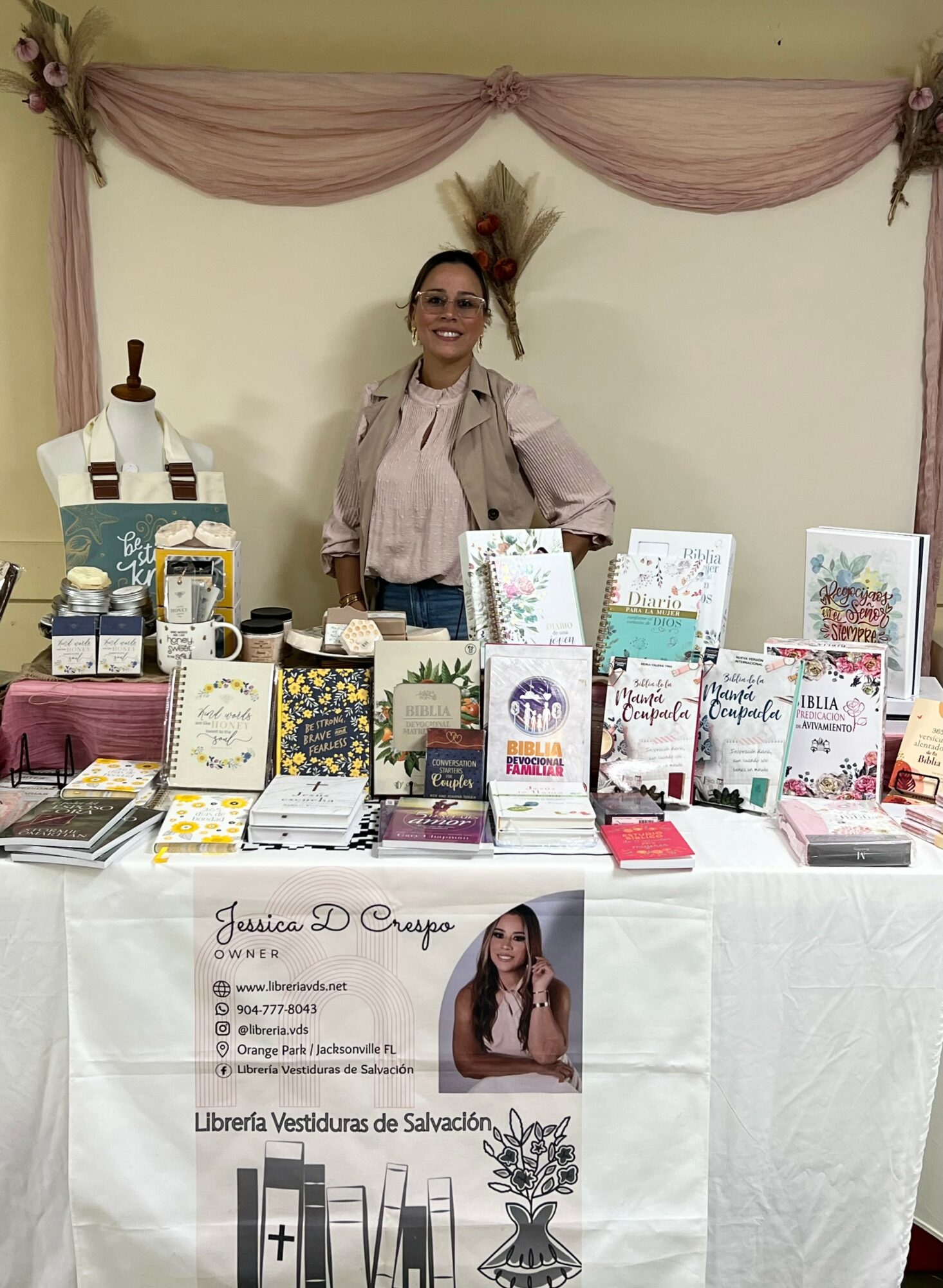 Woman standing behind a table with books and items, decorated with pink curtains and a wheat decoration, smiling.