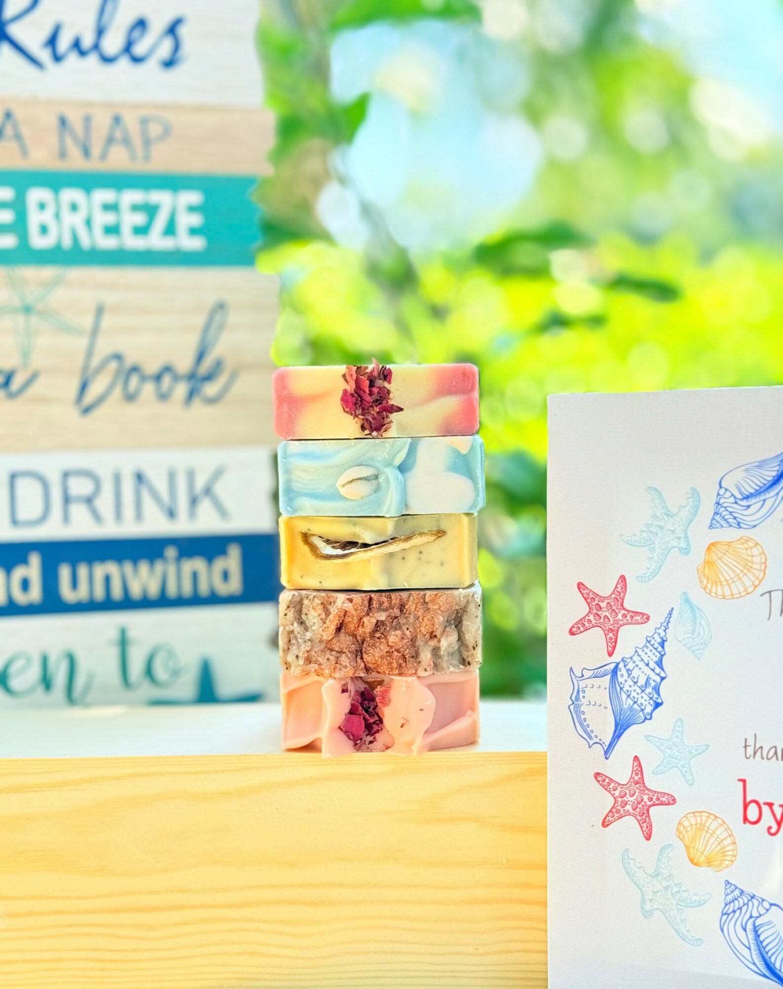Stacked colorful soap bars with ocean-themed drawing on paper beside a blurred outdoor background.