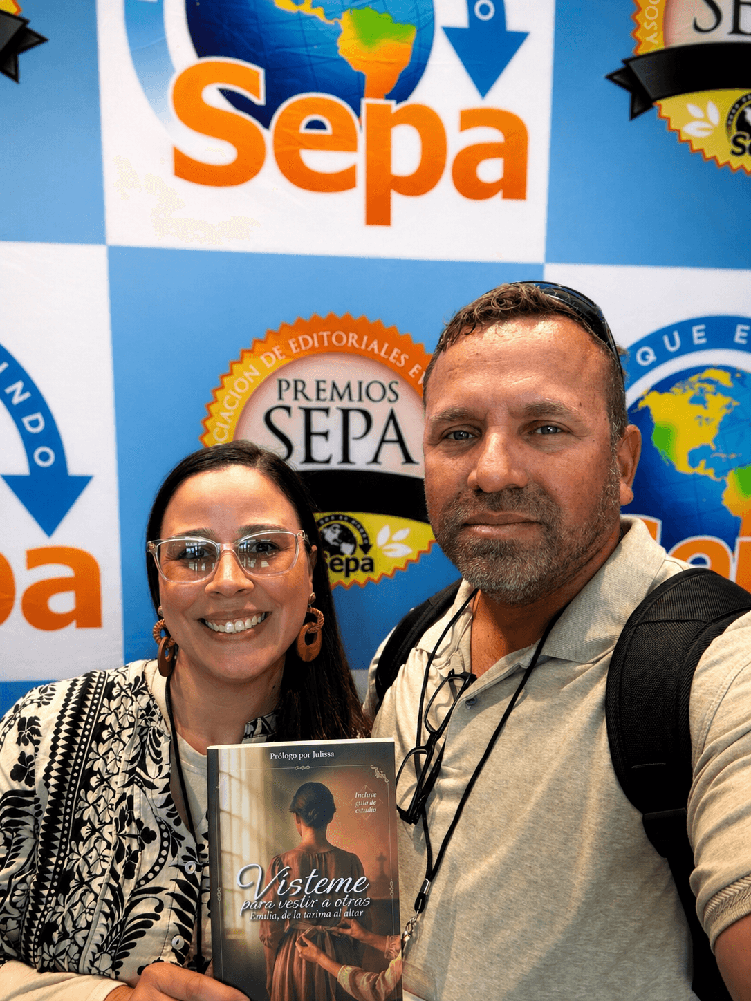 Two people pose in front of a blue backdrop with logos, one holding a book titled 'Vísteme'.