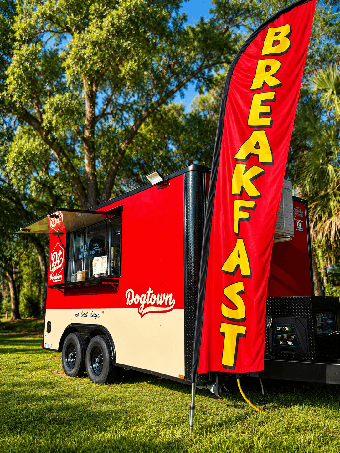 Food truck with a red and white exterior, a tall red flag with yellow text reading 'BREAKFAST', parked on grass with trees in background.