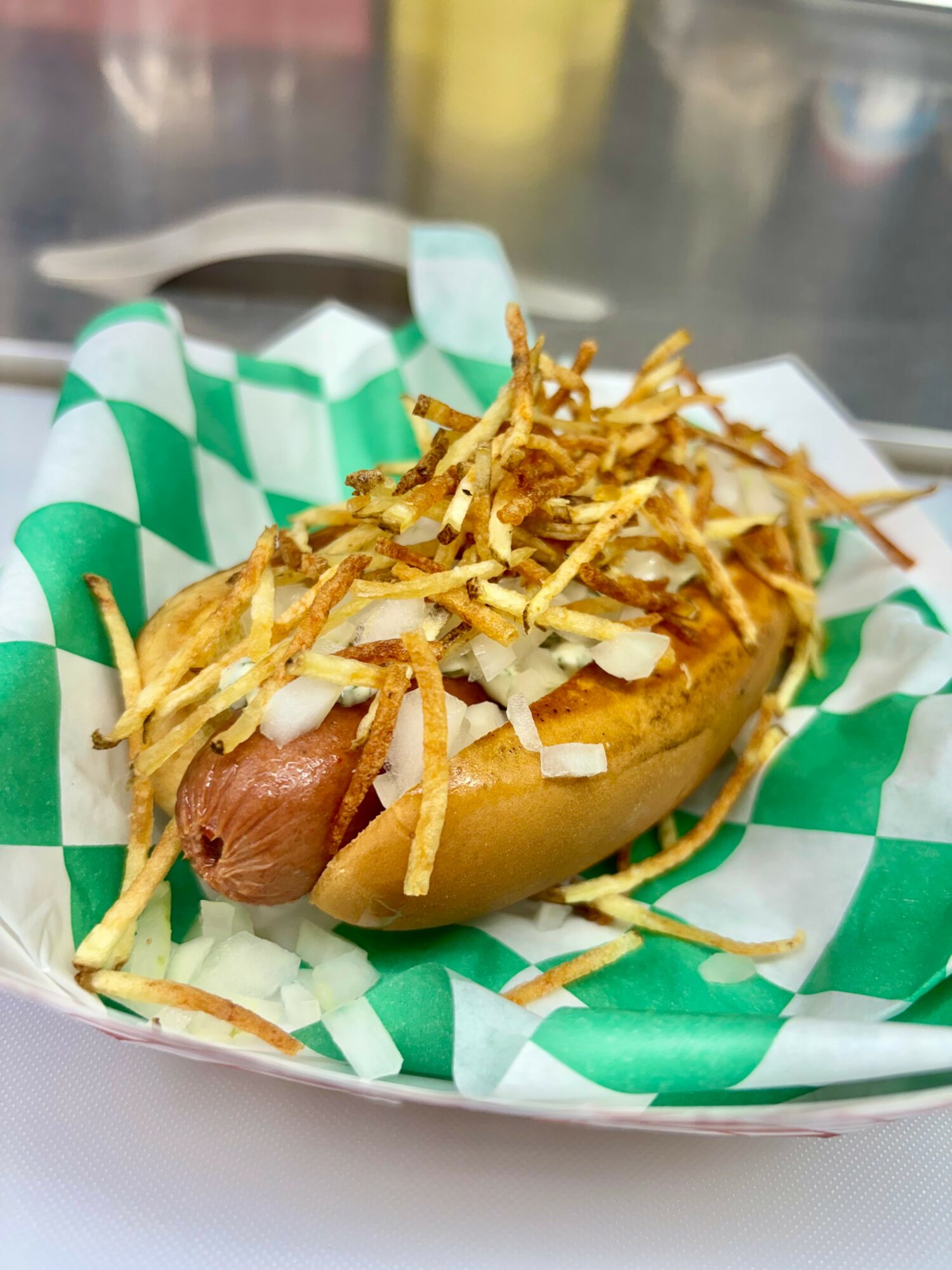 Hot dog with shredded cheese and onions in a bun on green and white checkered paper, in a basket.