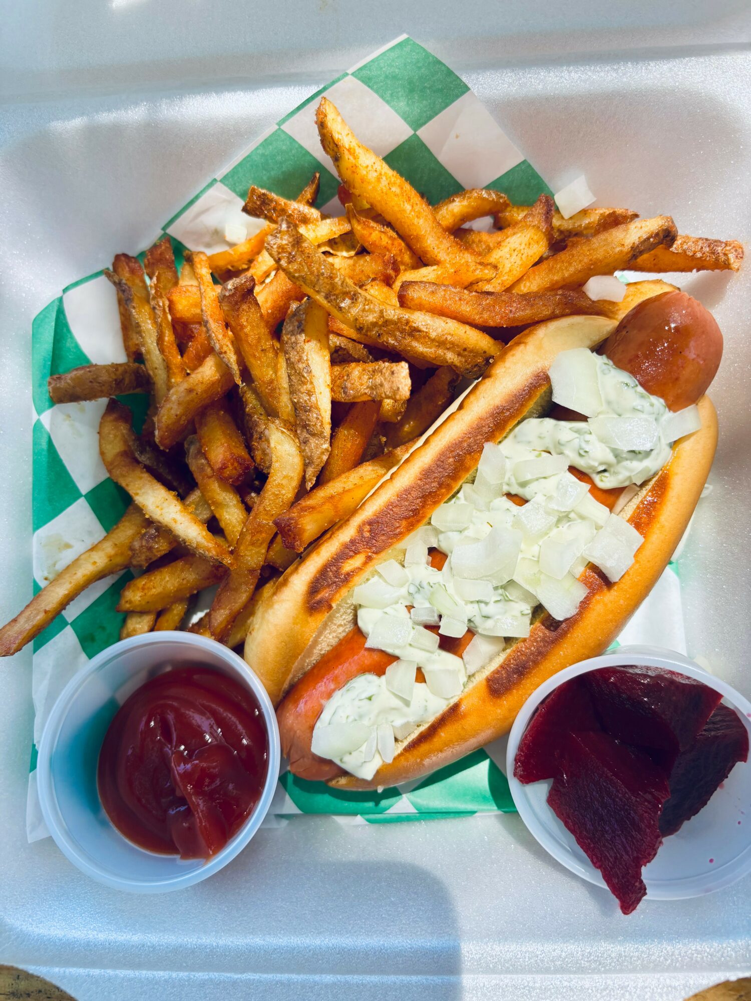 Hot dog with chopped onions in a bun, served with French fries and two cups of ketchup and barbecue sauce.