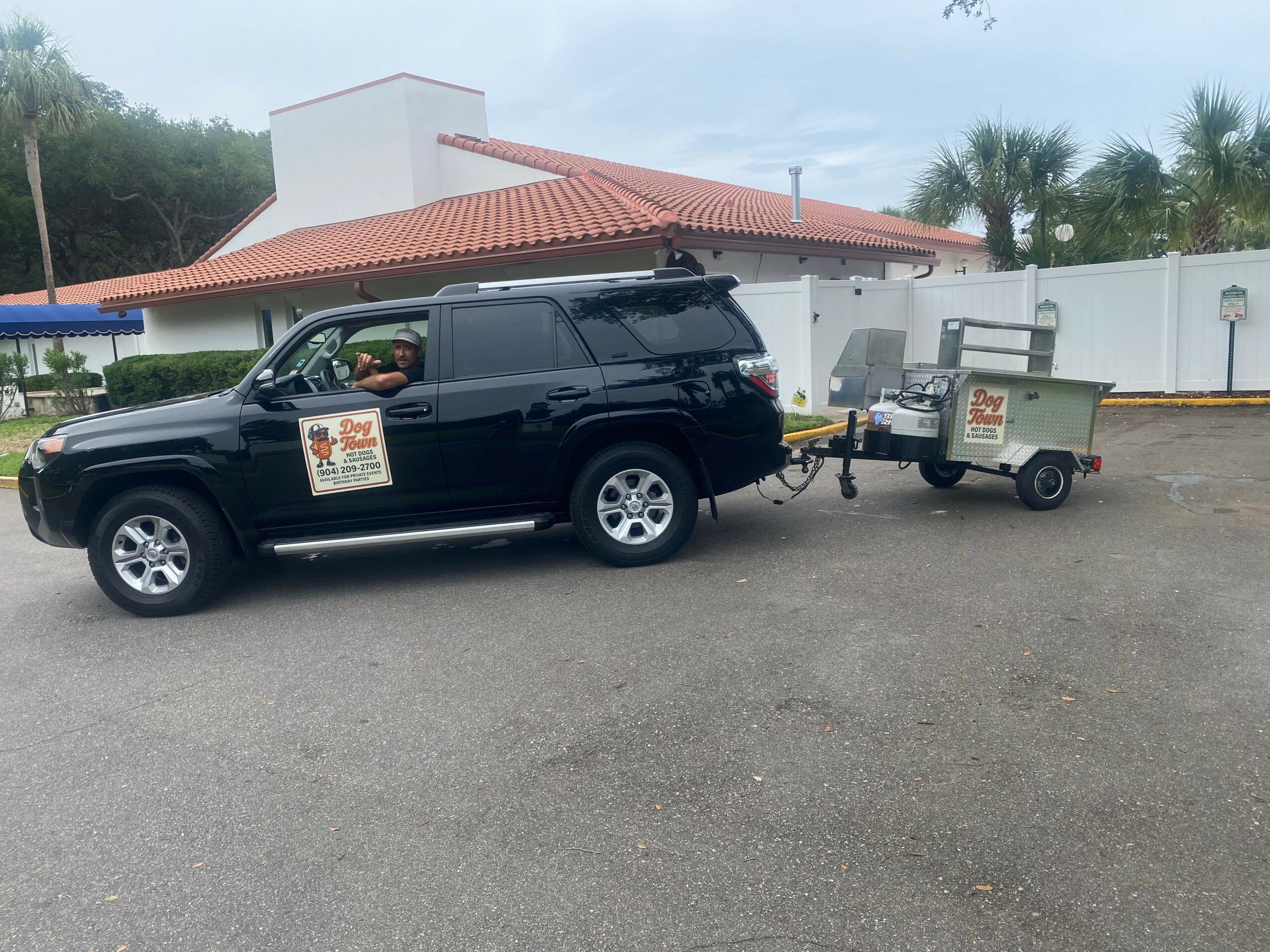 Black SUV with a trailer attached, parked outside a building with a red-tiled roof, trees, and a white wall in the background.