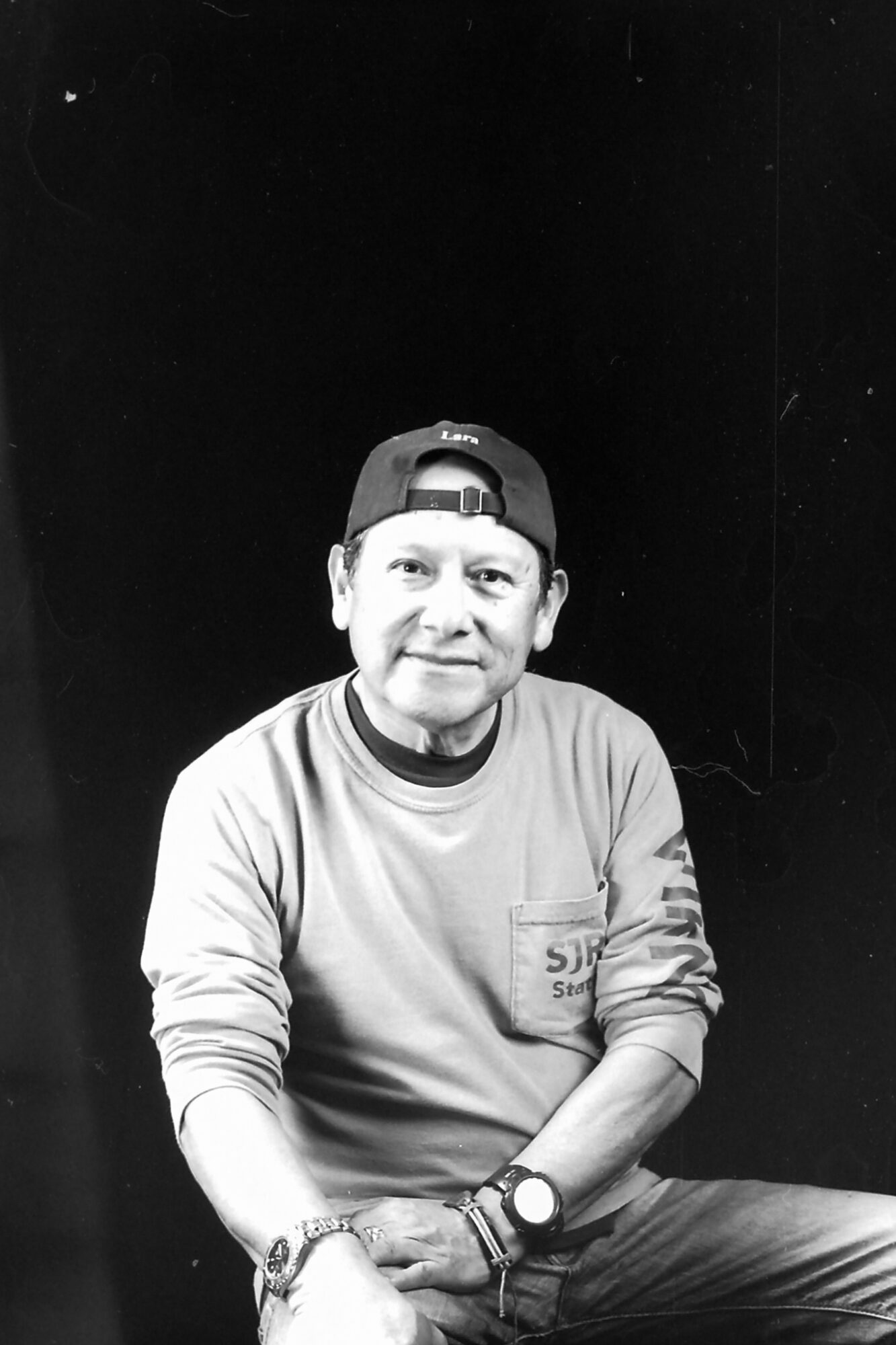 Person wearing a cap backward, sitting with arms crossed, smiling, against a black background.