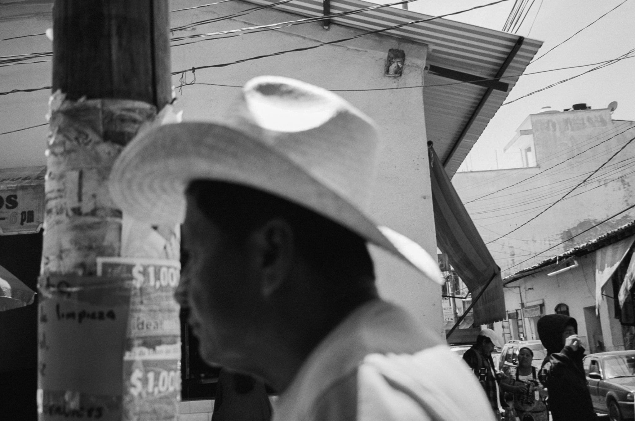 Silhouette of a person wearing a wide-brimmed hat in an urban street scene, with buildings and power lines in background.