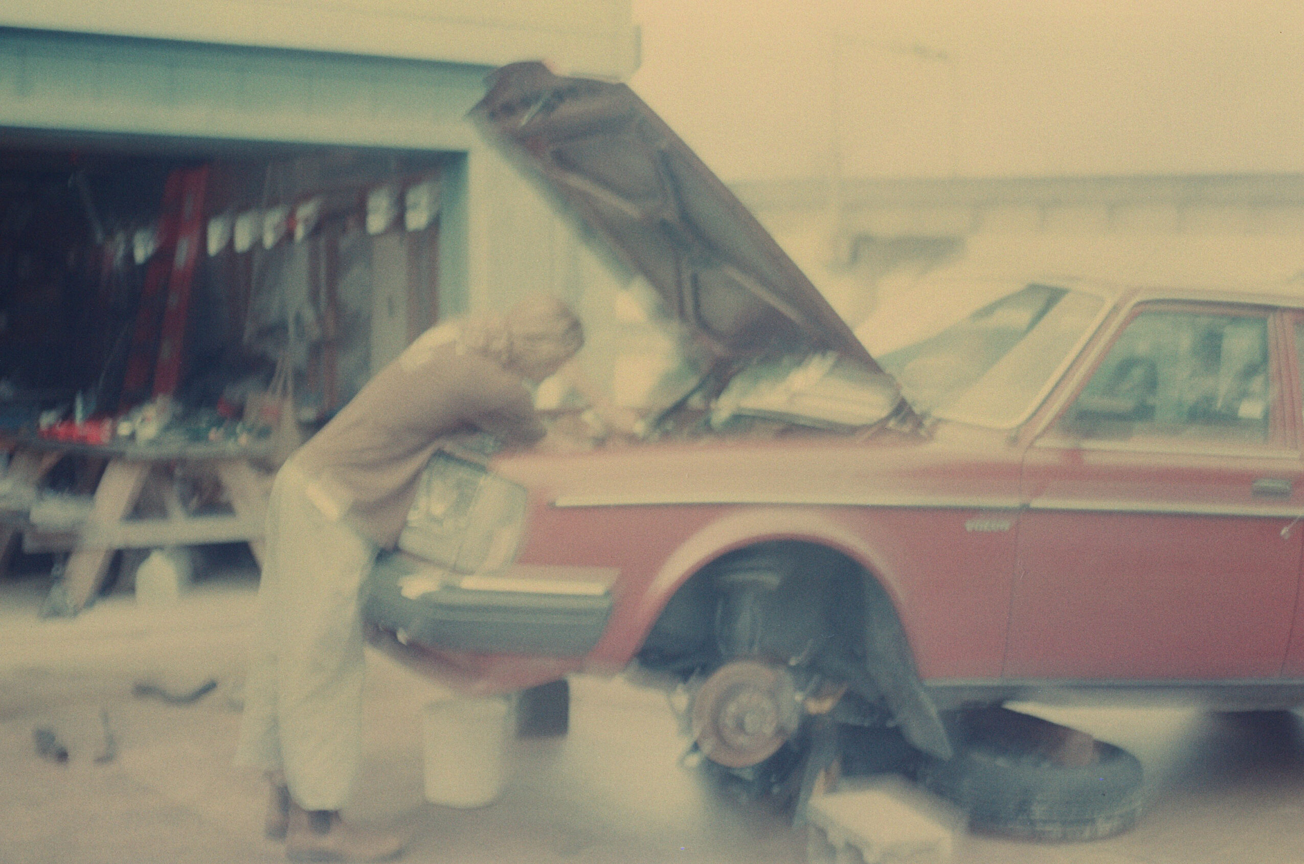 Person working on a car with the hood open, missing a wheel, in front of a building with a red and white exterior.