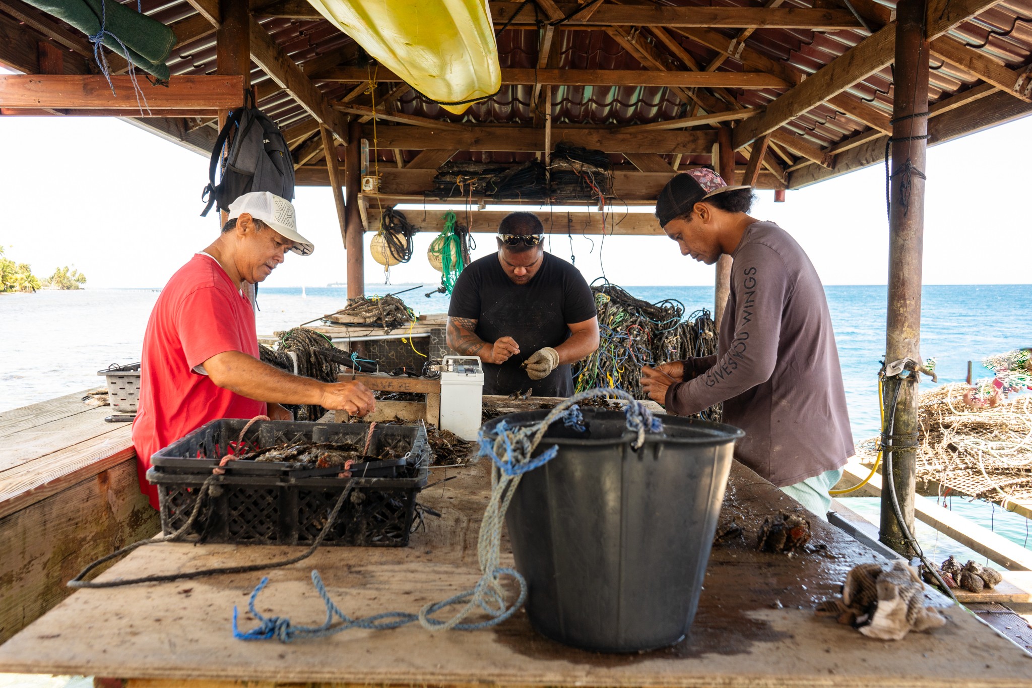 Three people working at a beachside wooden shelter, handling fishing equipment with water and boats in the background.