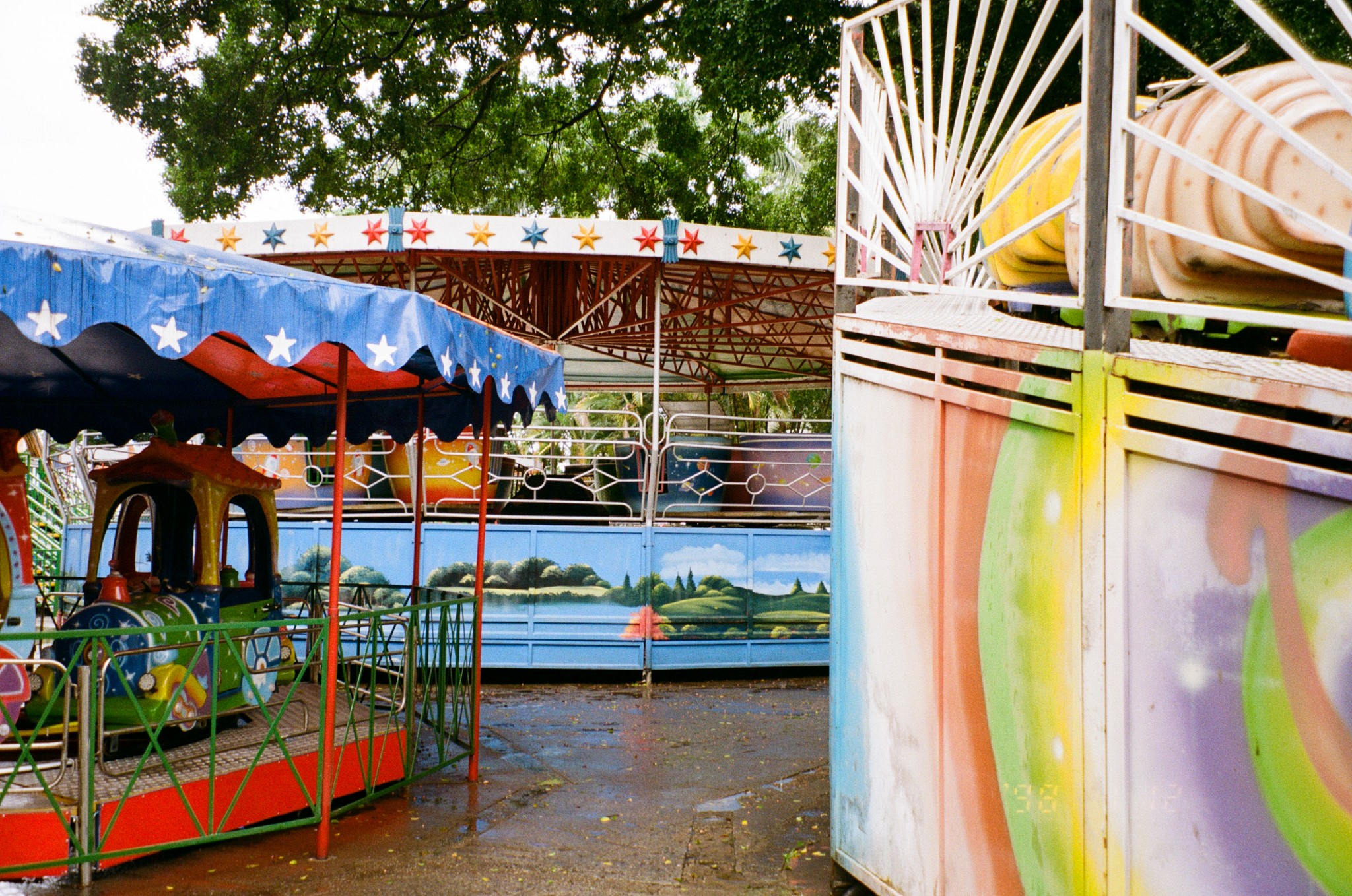 Colorful amusement park rides with a carousel and a spinning ride, under a tree, with bright decorations and a cloudy sky.