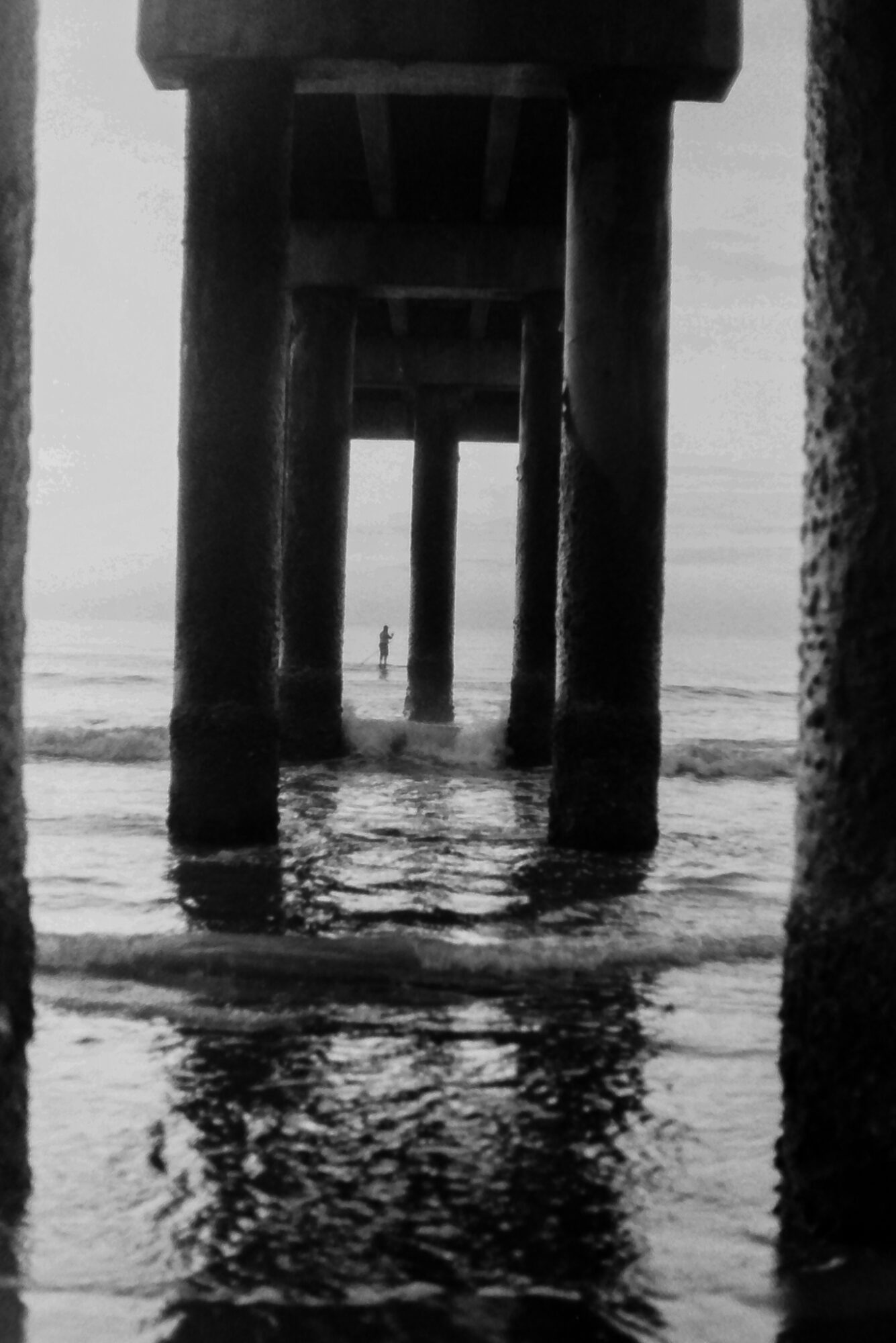 View under a pier with large supporting columns extending into the water, a person standing in the distance, water reflections visible.