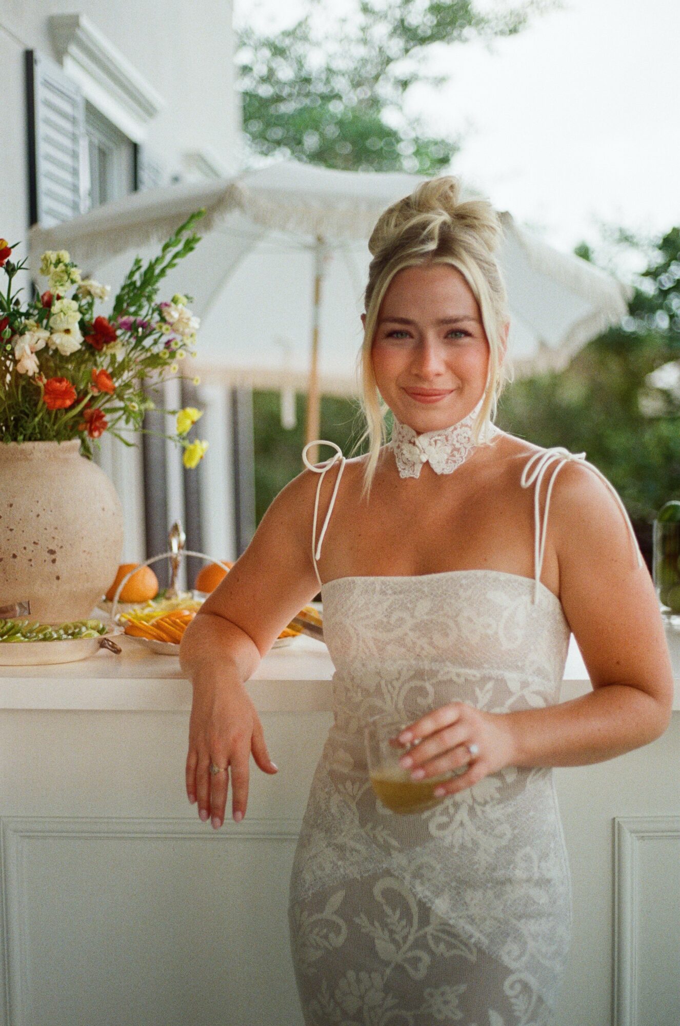 Woman in wedding dress holding a glass, smiling, with flowers and greenery in background.