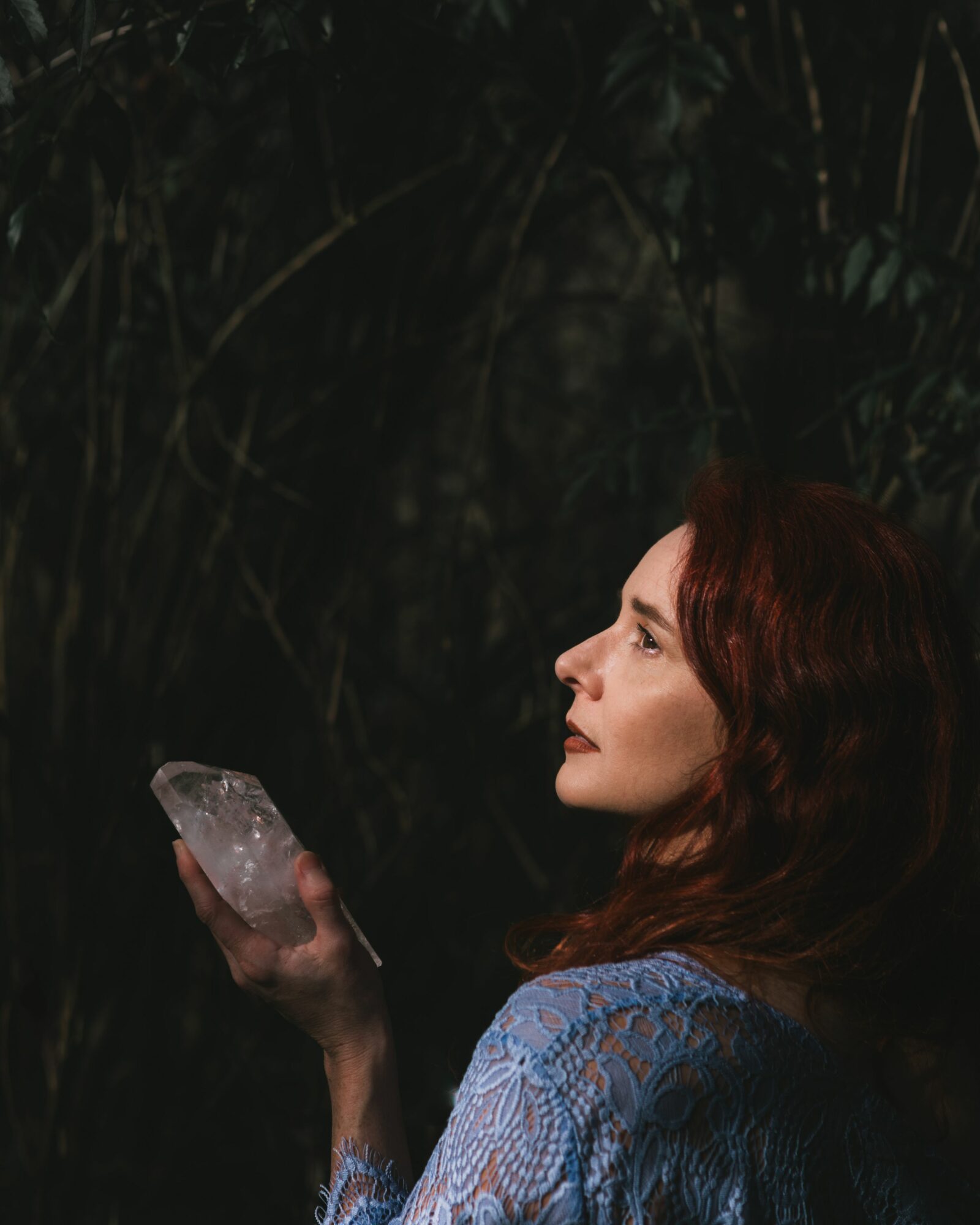 Woman with red hair in profile holding a large clear crystal against a dark background.