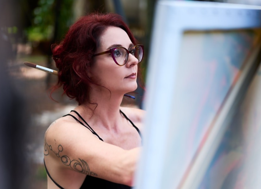 Woman with red hair and glasses painting outdoors, with trees in the background.
