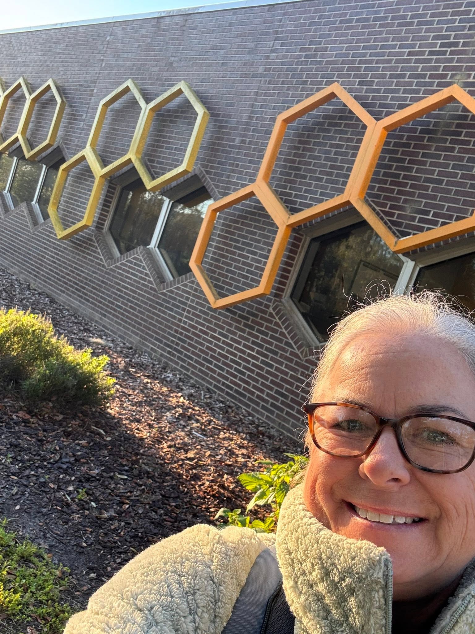 Person with glasses smiling outdoors in front of a brick wall with honeycomb-shaped frames.