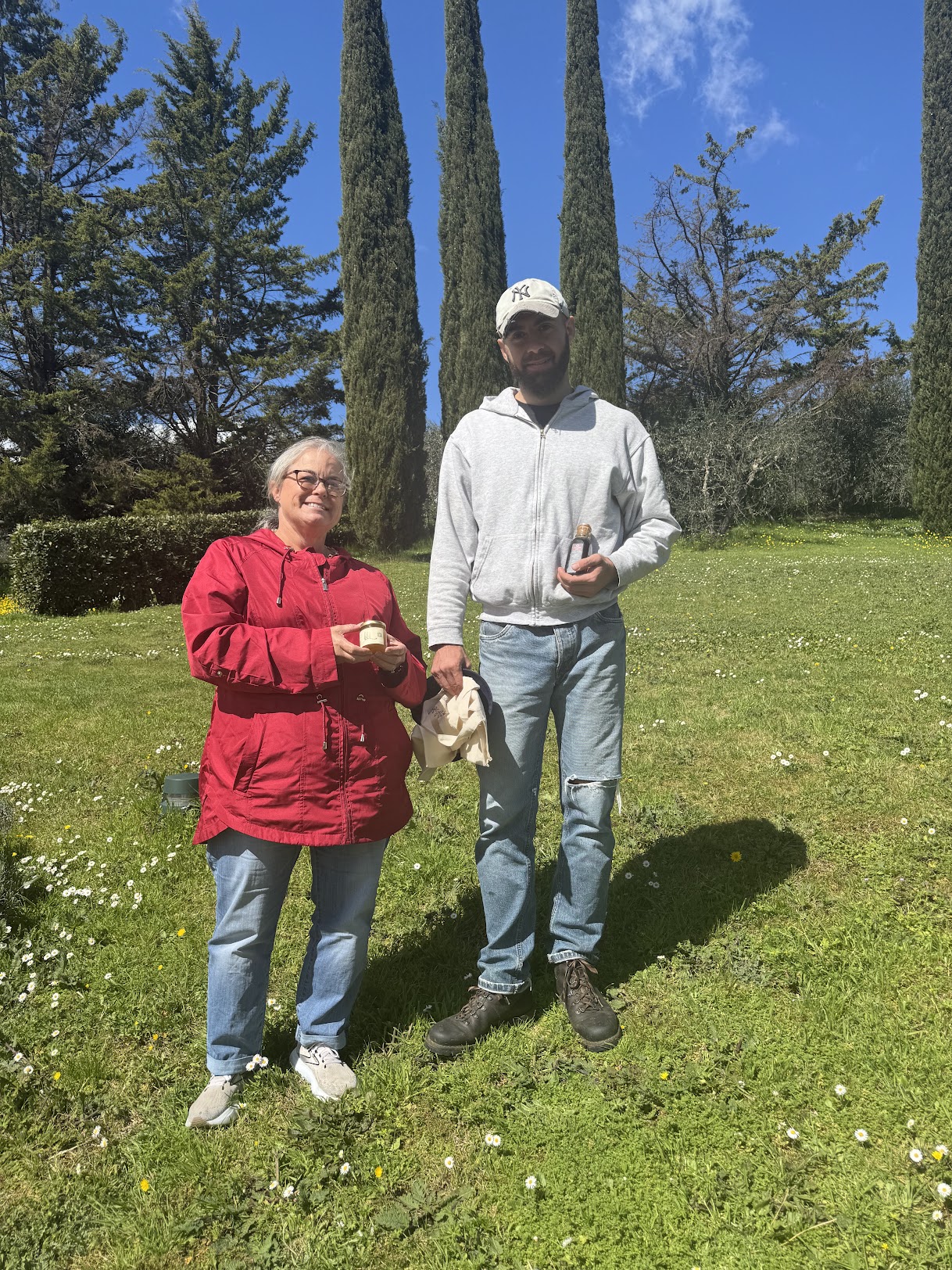 Two people standing outdoors on grass with trees and blue sky in background, holding objects, smiling, sunny day.