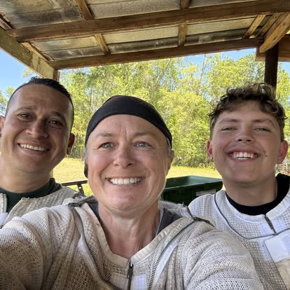 Three people smiling outdoors under a wooden shelter with trees in the background.