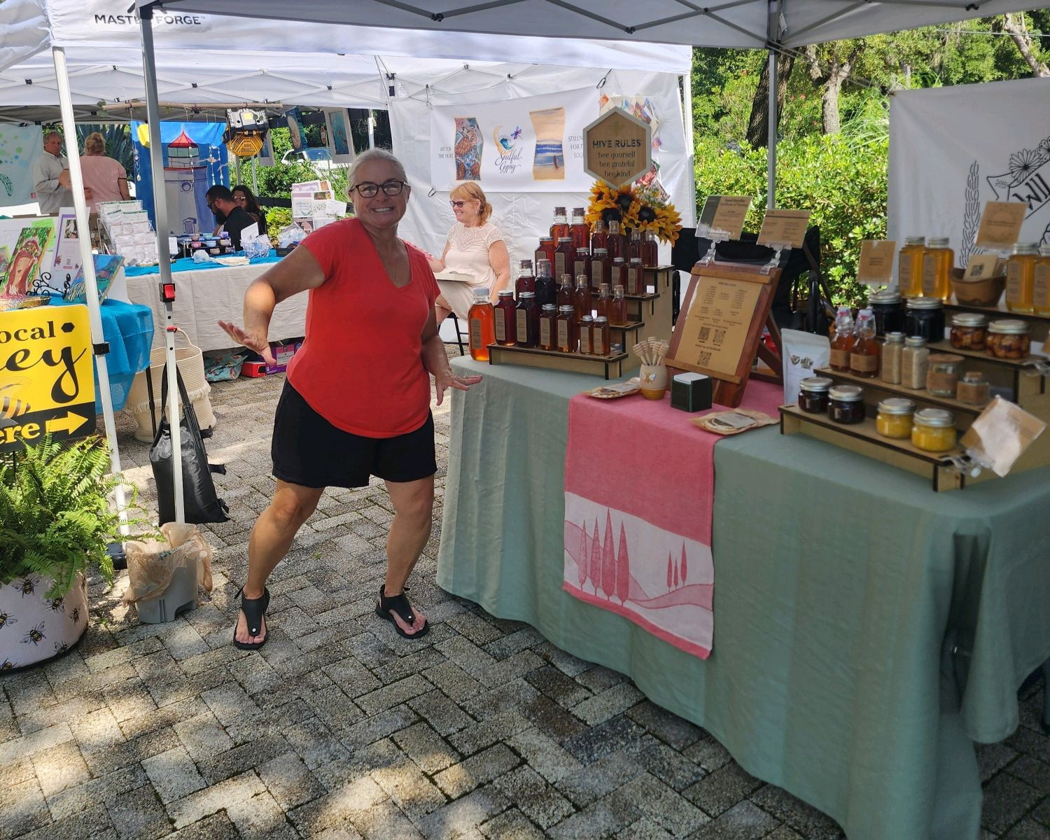 Person standing at outdoor market stall with jars and bottles, tents, and other vendors in background.