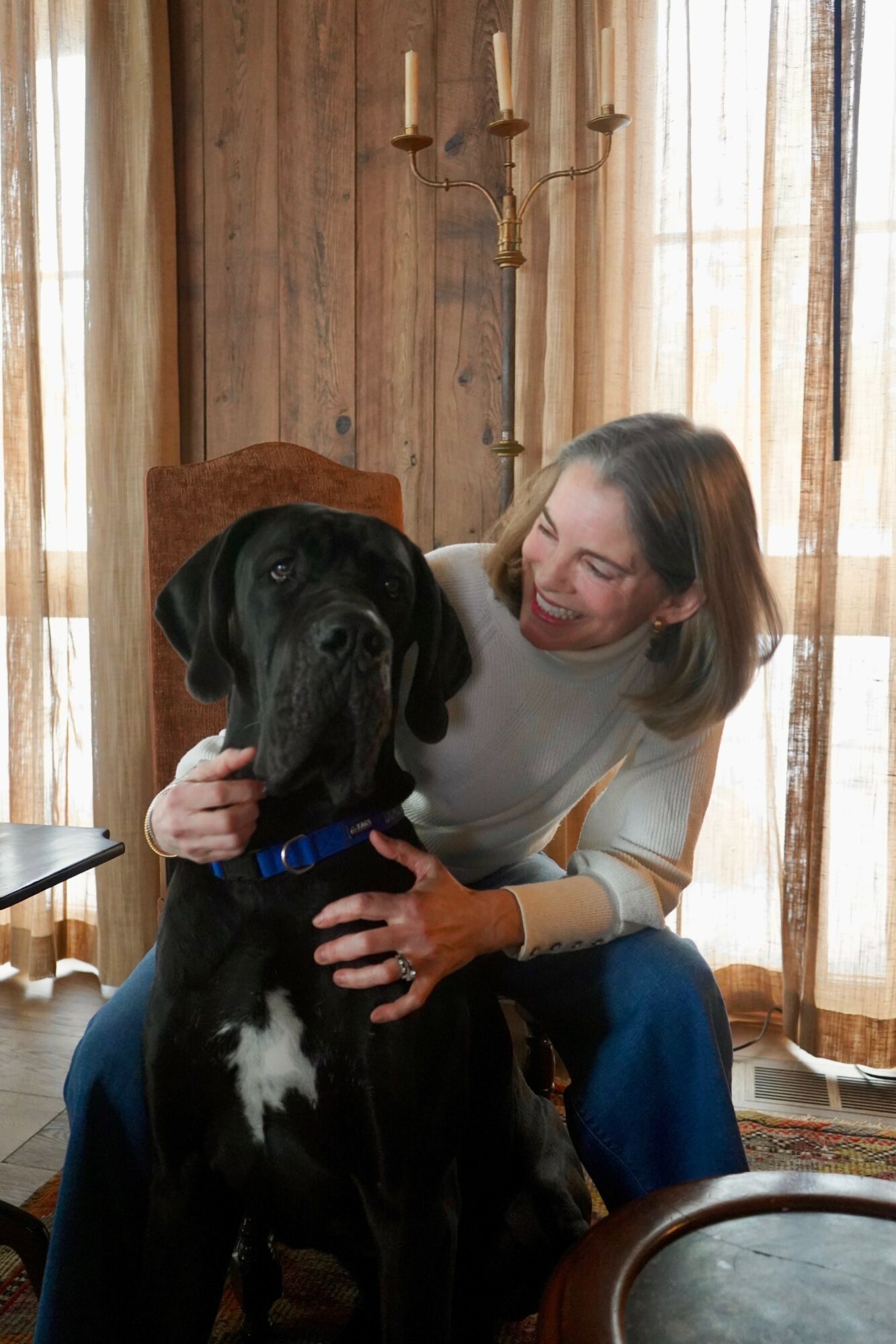 Woman smiling and hugging a large black dog indoors with wooden walls and curtains behind.