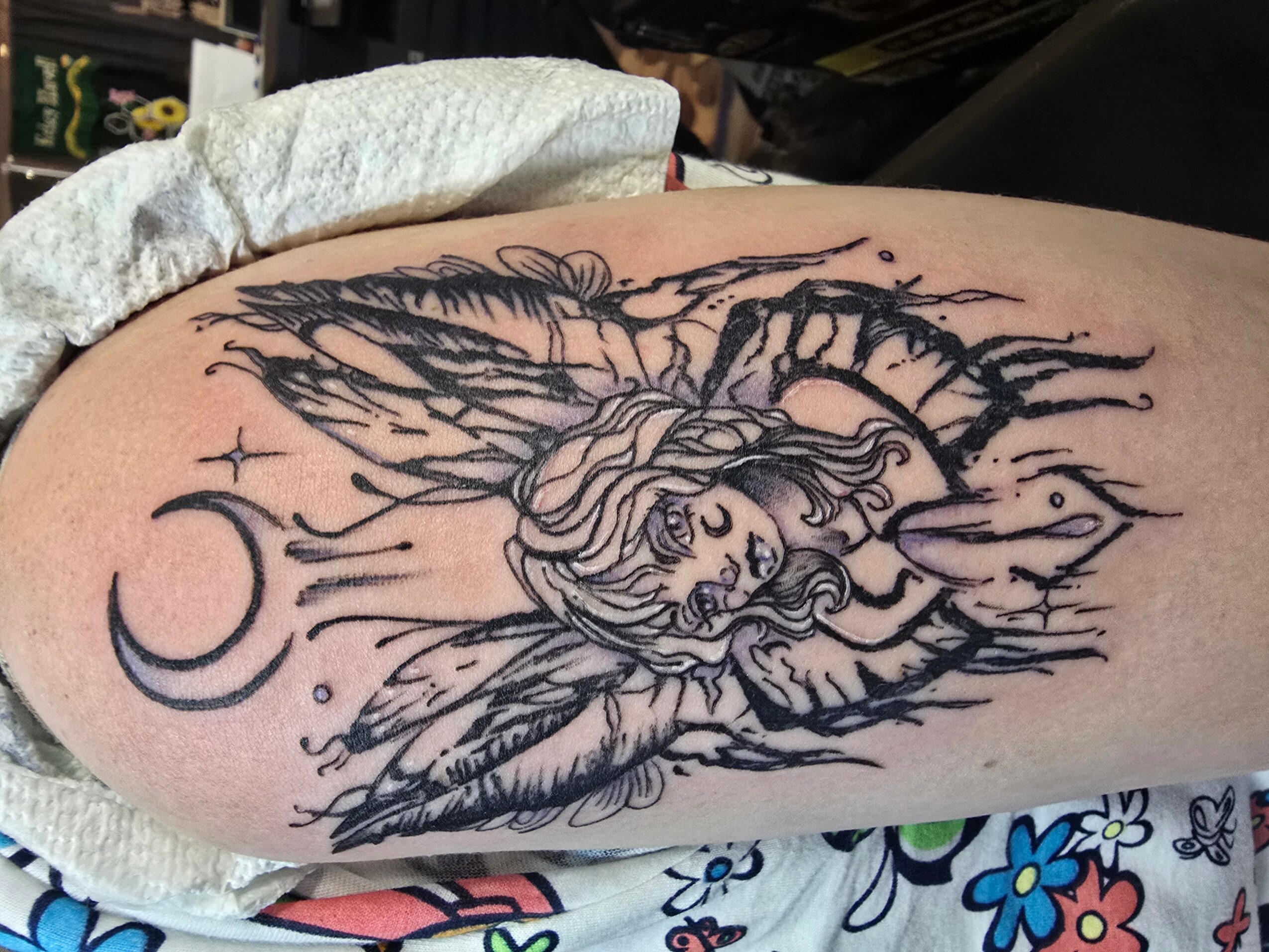Tattoo of a fairy with wings and flowing hair, surrounded by moon and stars, on person's arm.