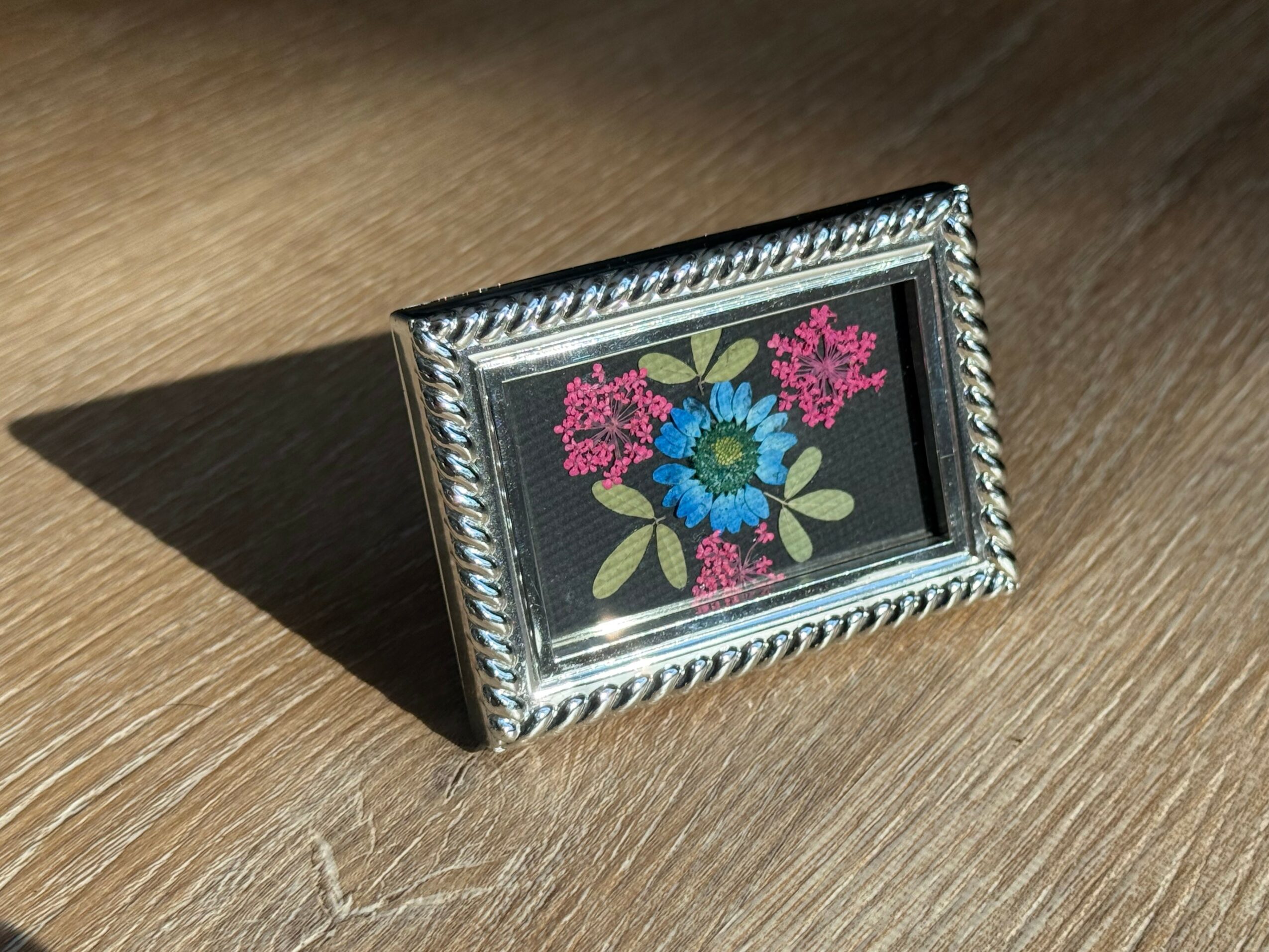 Small framed floral embroidery with pink, blue, and green flowers on a black background, casting a shadow on a wooden surface.