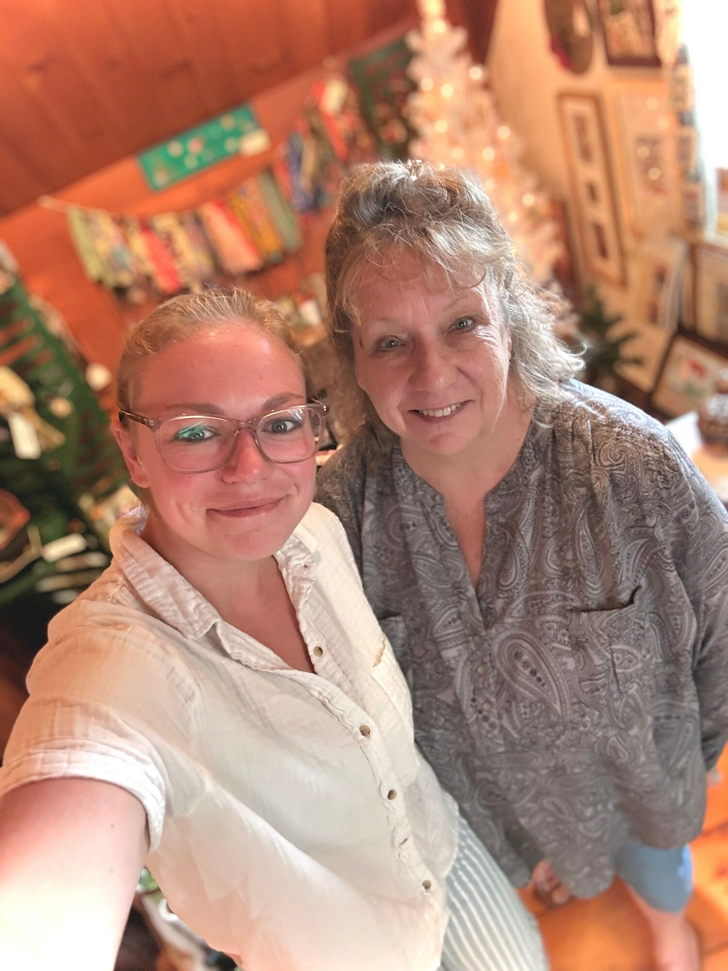 Two women smiling indoors with Christmas decorations and a Christmas tree in the background.