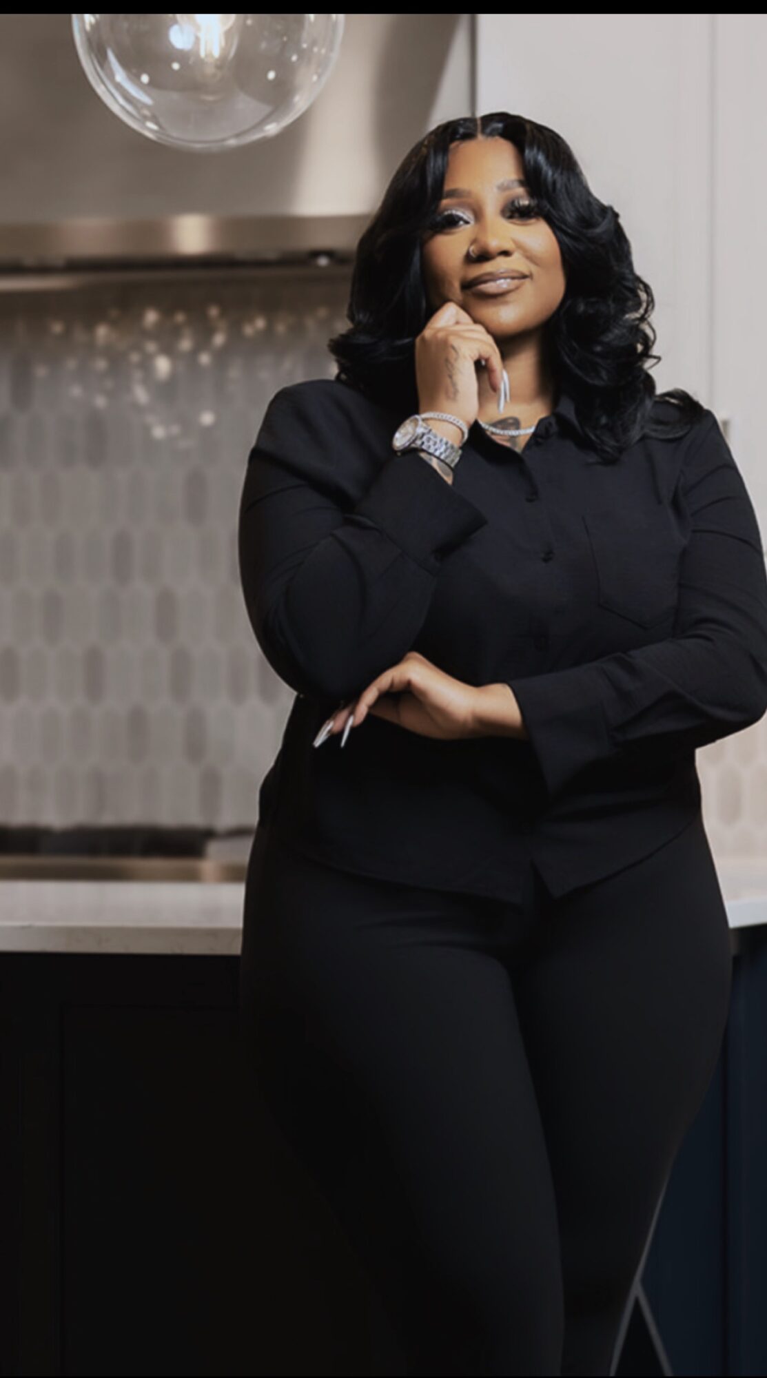 Woman with black hair in a black outfit, smiling, standing indoors with a mirror and decorative background.