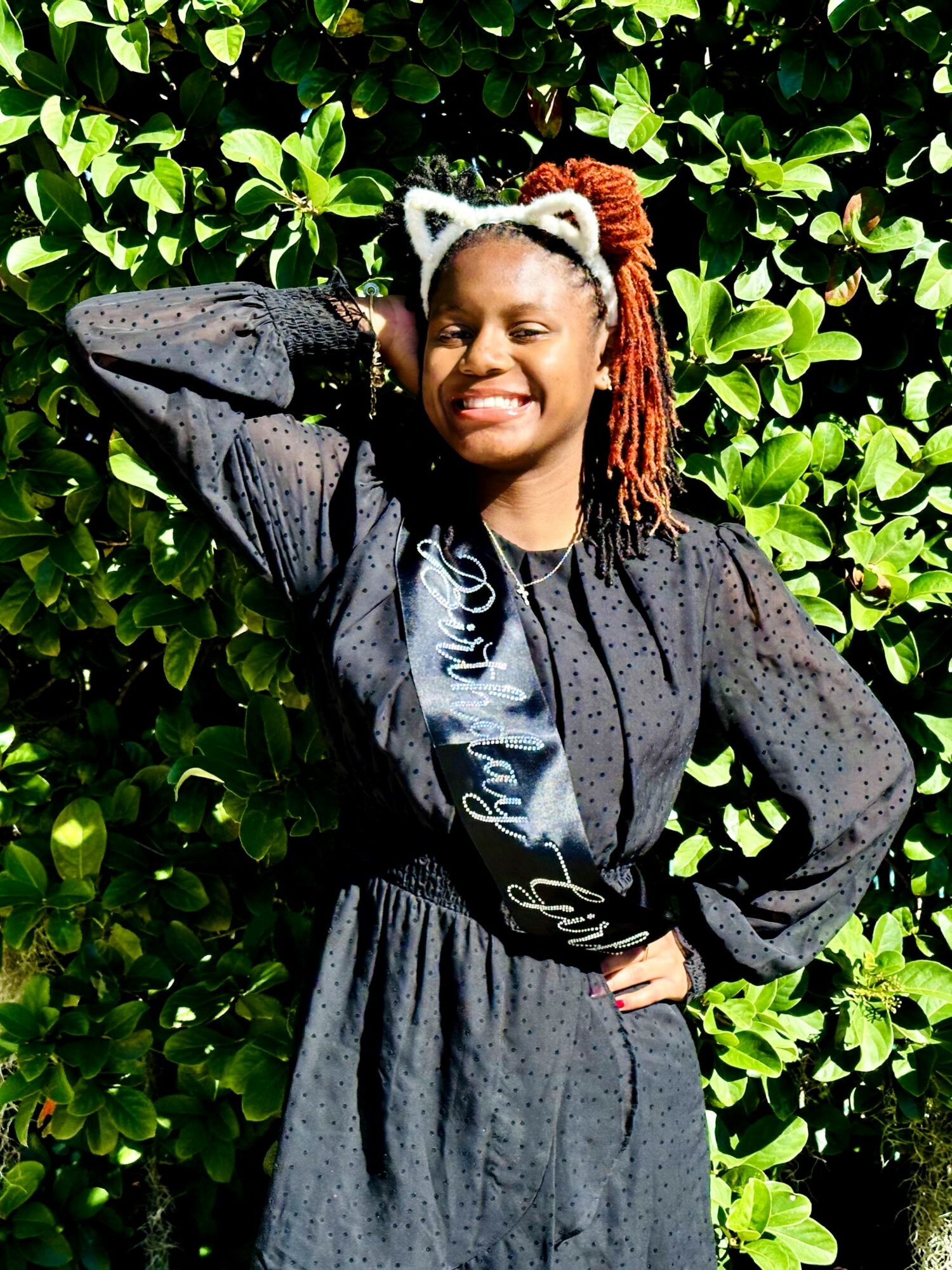 Smiling woman with braided hair and cat ear headband standing in front of green leafy background.