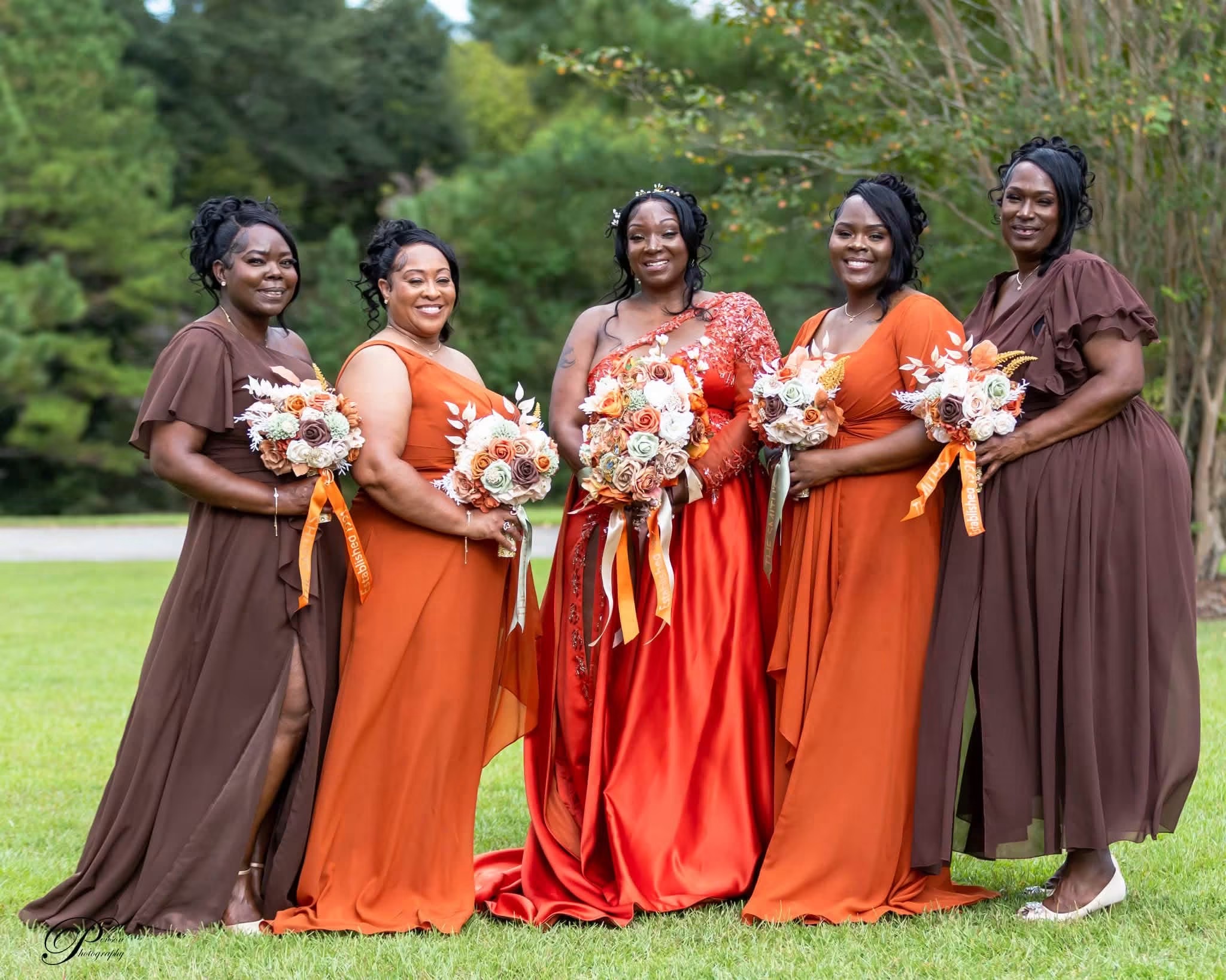 Five women in colorful dresses holding bouquets stand outdoors on grass with trees in background.