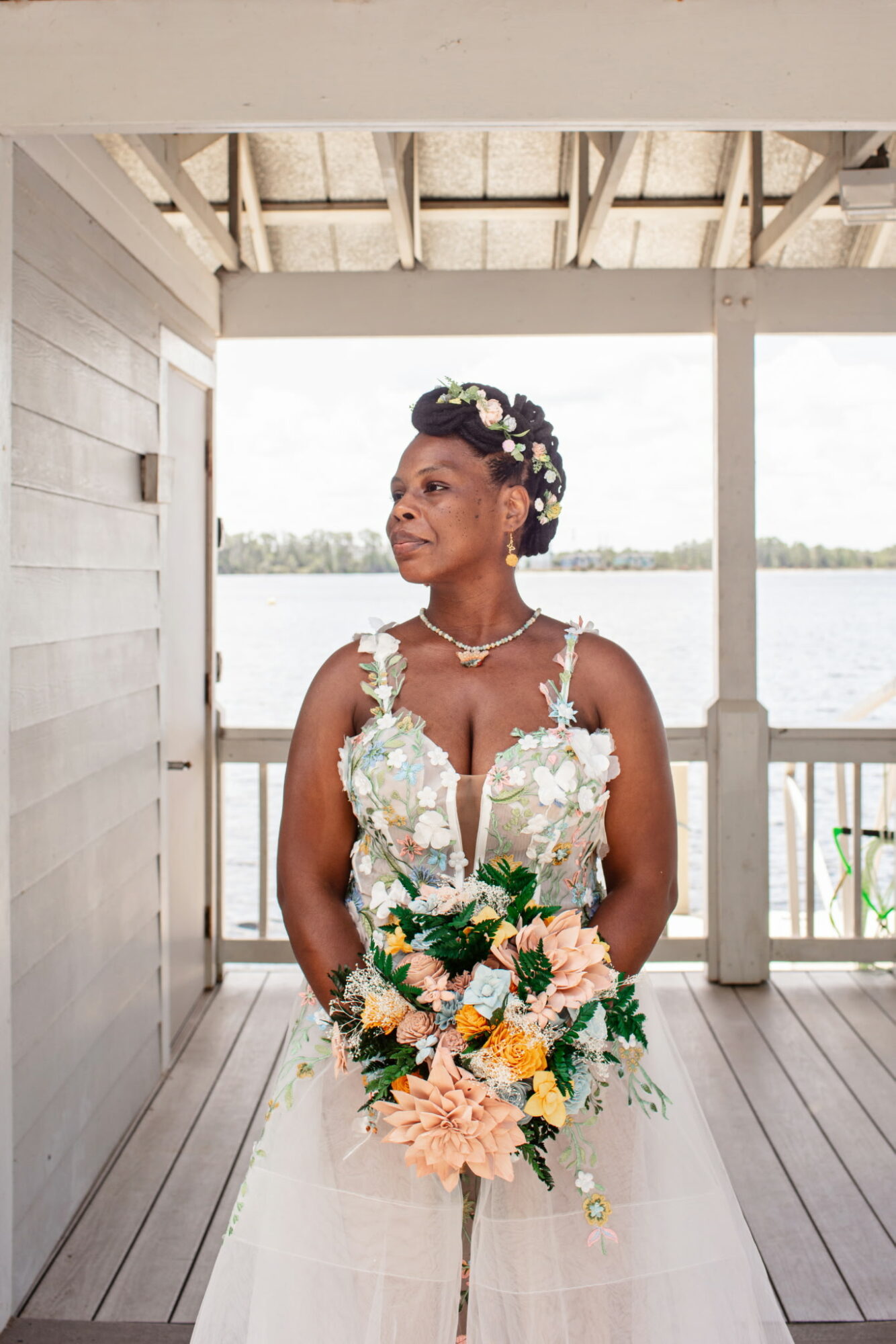 Woman in wedding dress holding a bouquet, standing on a wooden porch with water in the background.