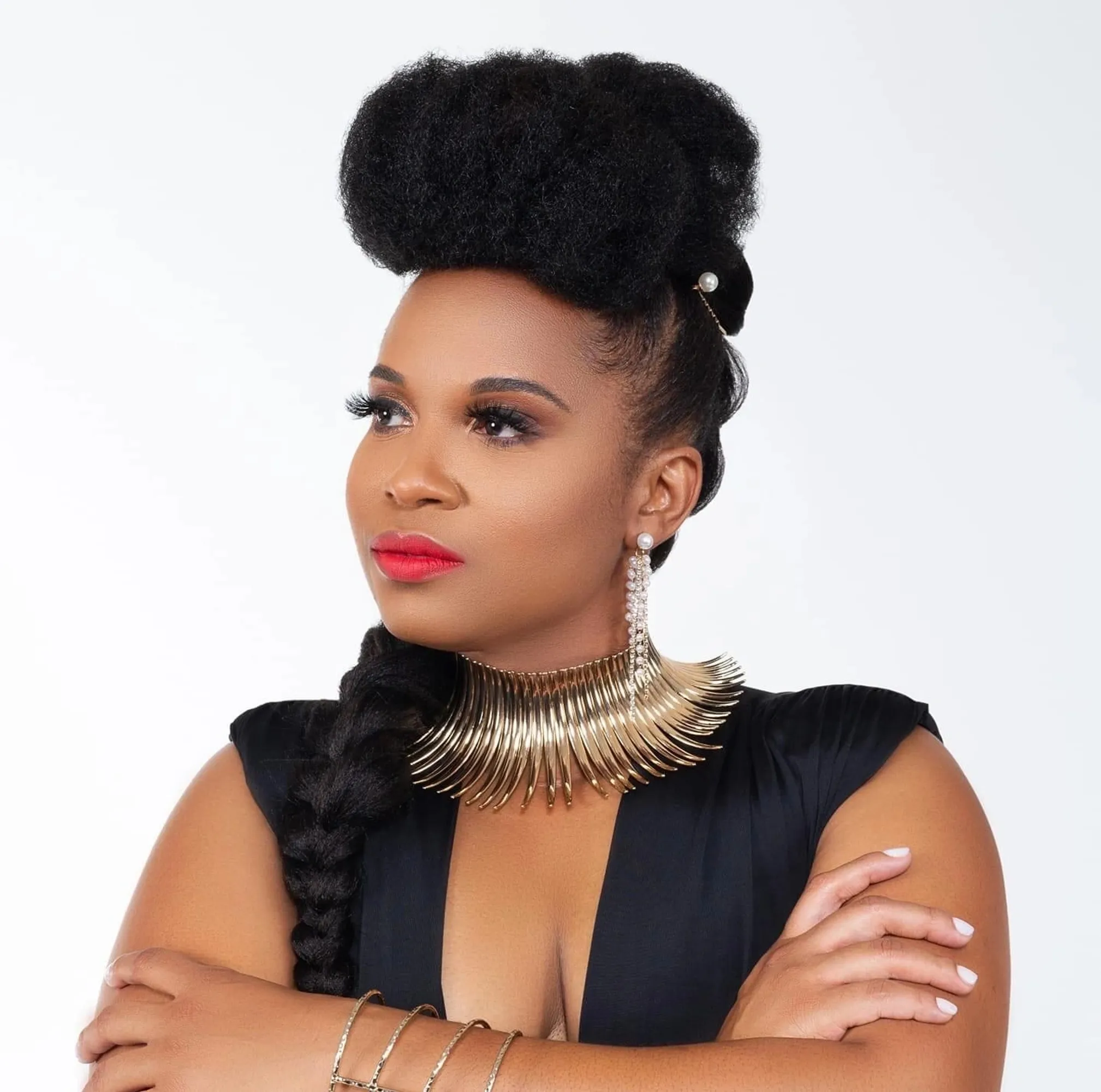 Woman with styled hair, wearing a black dress and large necklace, looking to the side with crossed arms.