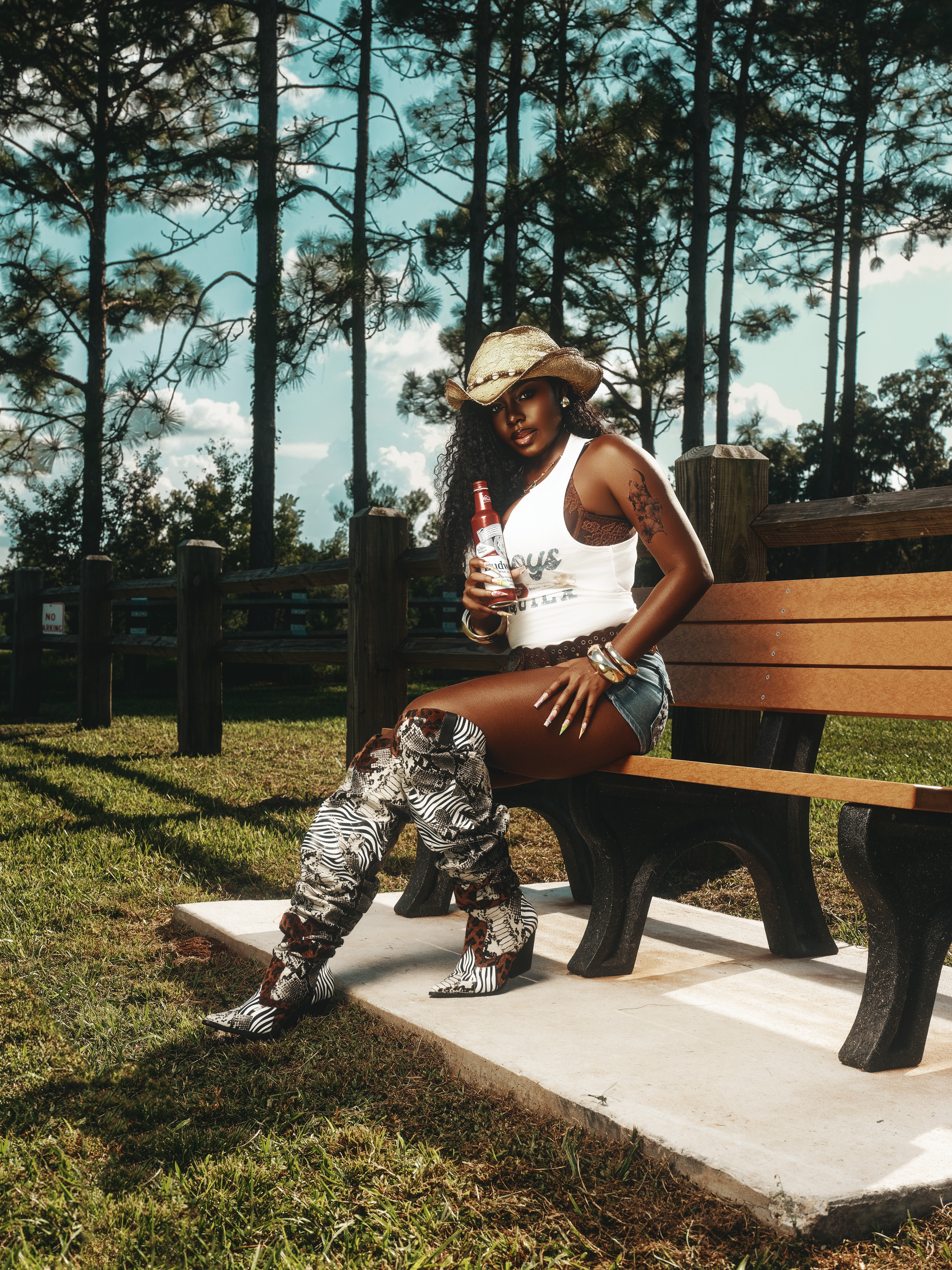 Woman sitting on a park bench holding a drink, wearing a hat, sunglasses, and casual clothing, with trees in the background.
