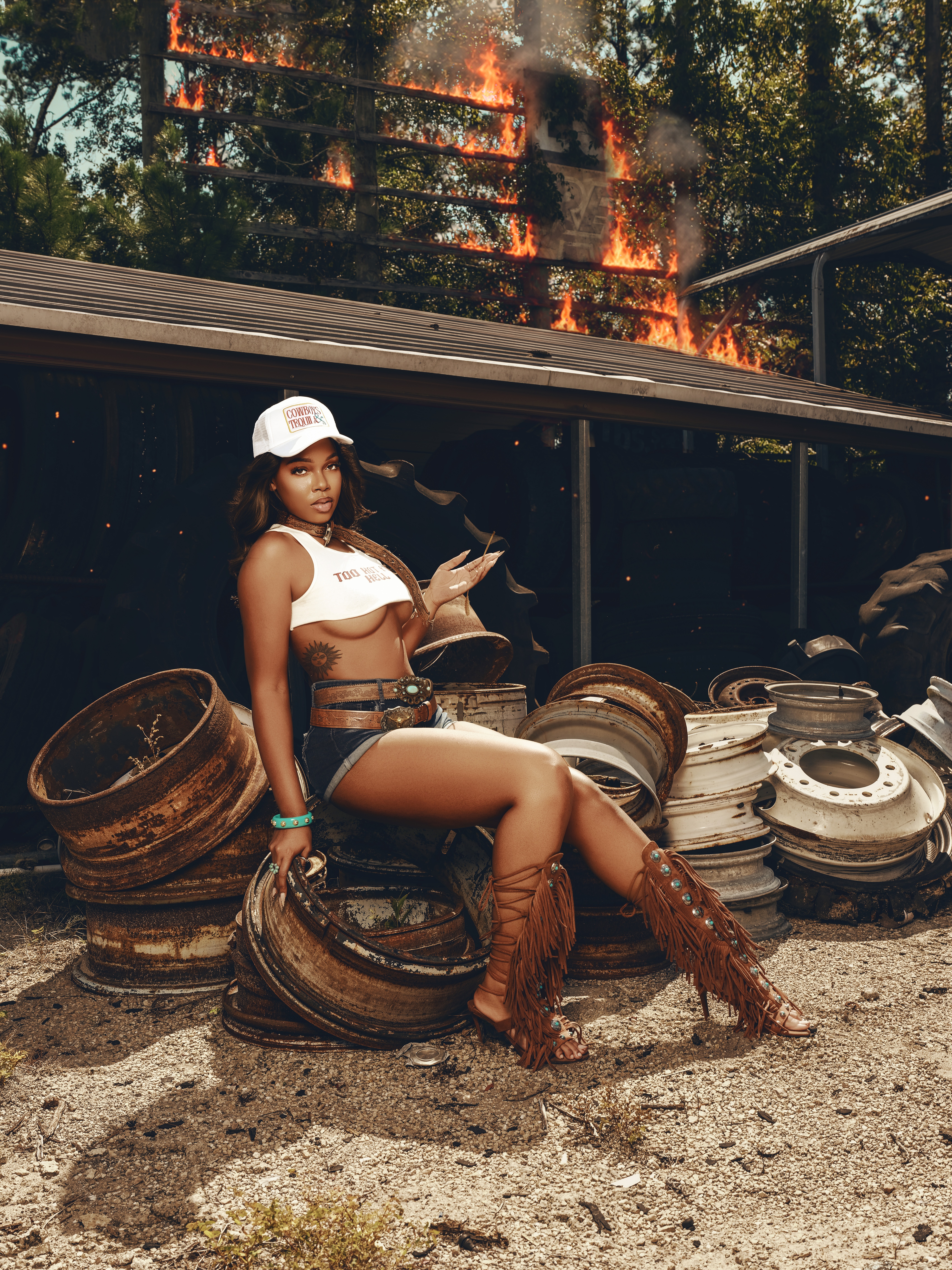 Woman in a white crop top, shorts, and boots sitting on metal drums outdoors with a fire burning behind her.