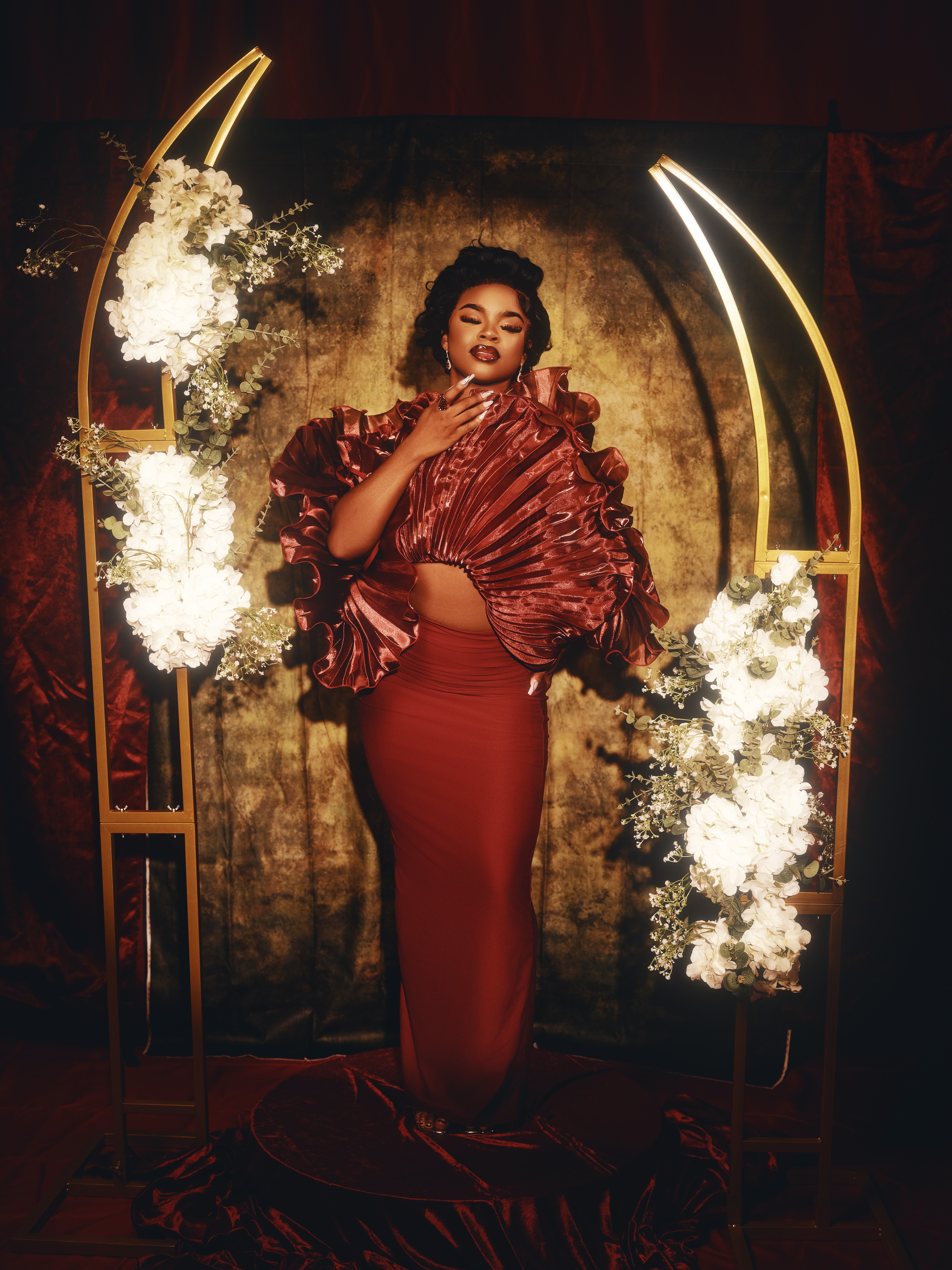 Woman in elegant red dress and ruffled top stands on stage with floral decorations and golden arch, posing confidently.