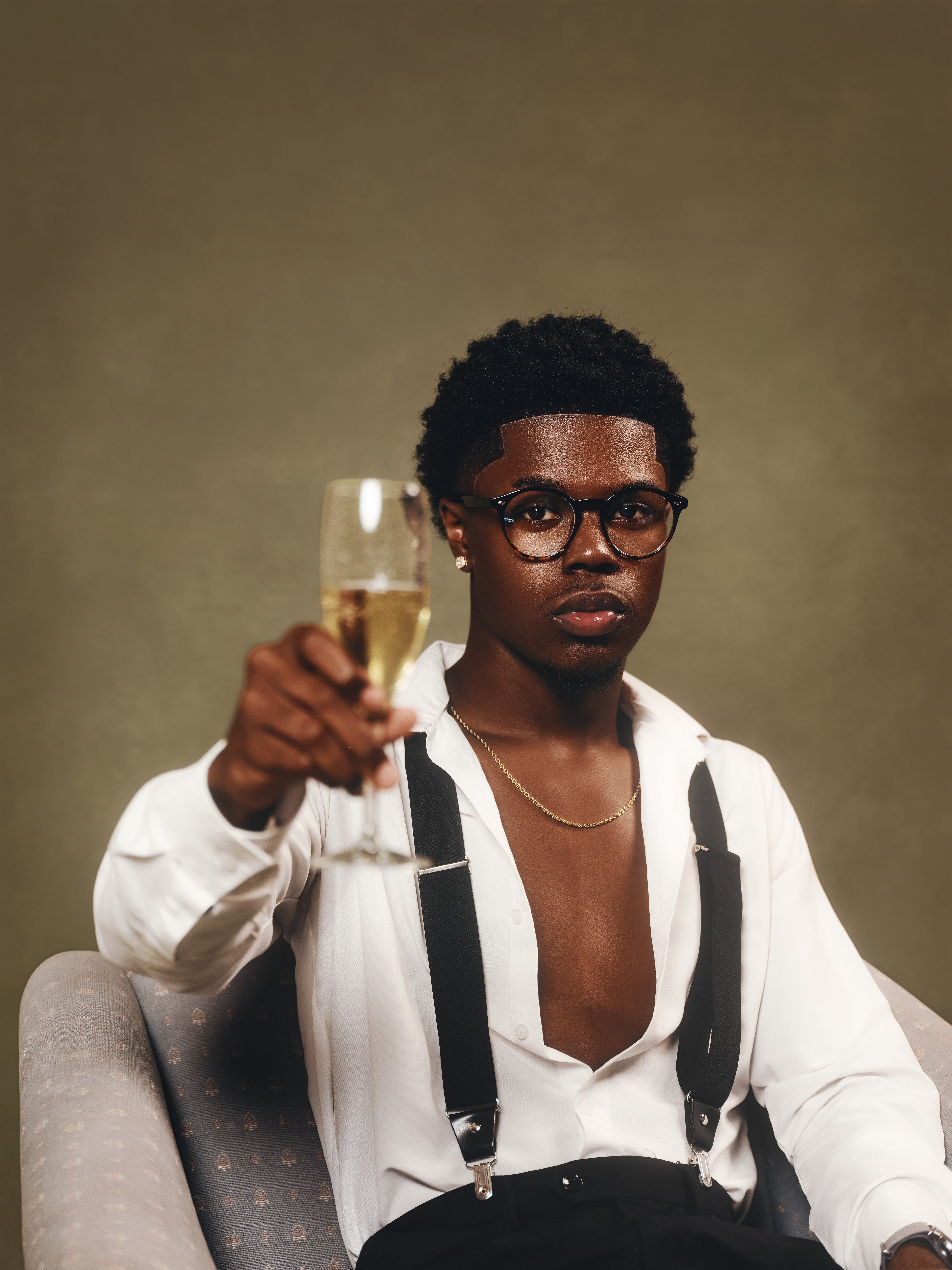 Person with short curly hair and glasses holding a glass of champagne, wearing a white shirt with suspenders, against a plain background.