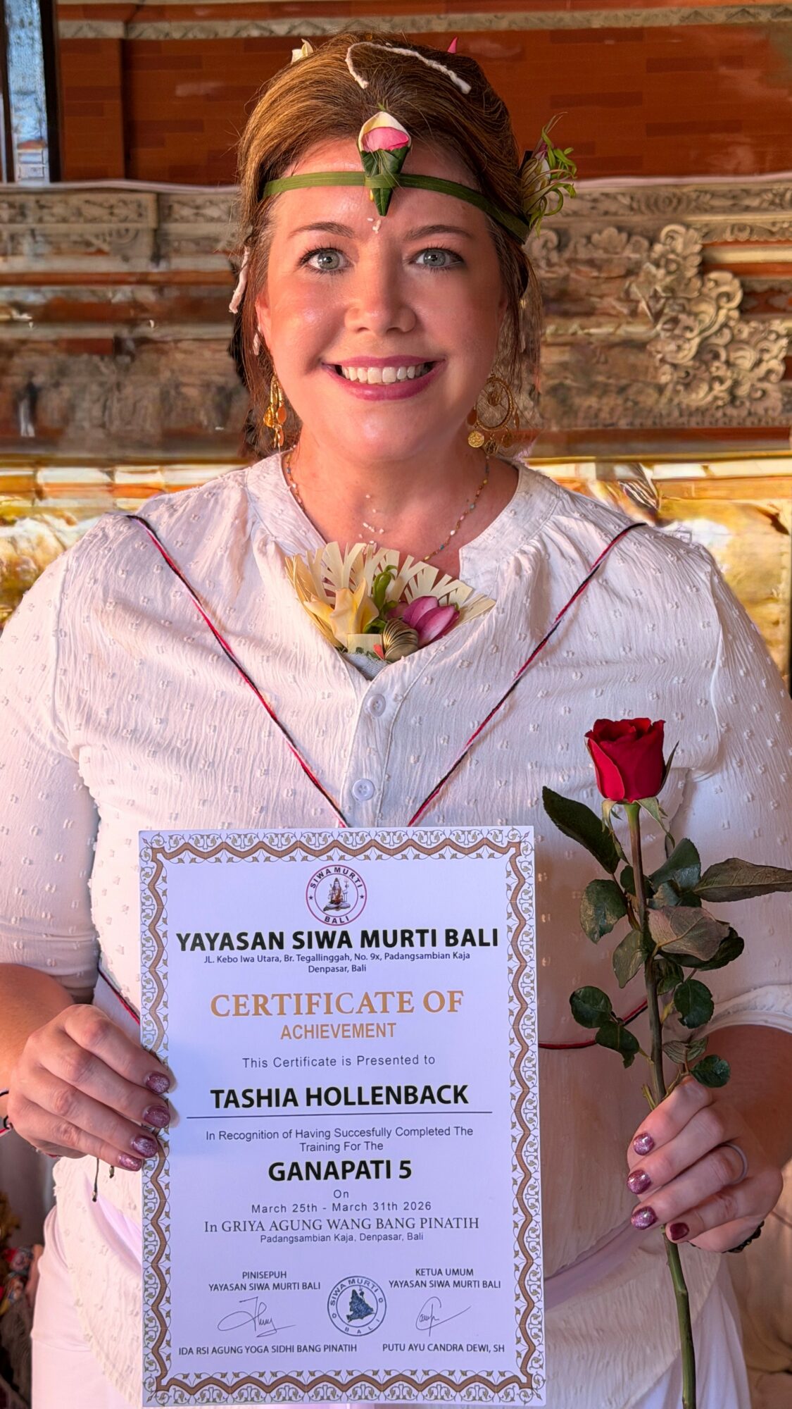 Smiling woman in white dress holding a red rose and certificate, with a floral headband and background of wooden decor.
