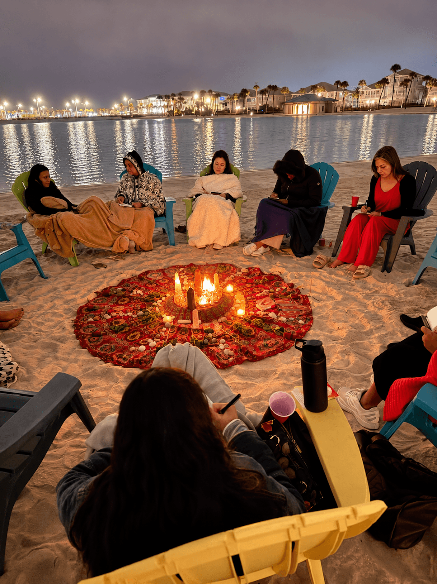 Group of people sitting on chairs around a fire pit on a beach at night, with city lights reflecting on water in background.