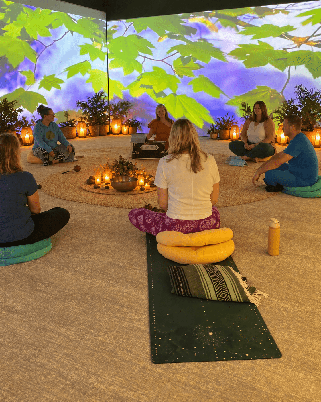 Group of people sitting in a circle on cushions and mats, participating in a meditation or yoga session indoors with a nature-themed backdrop.