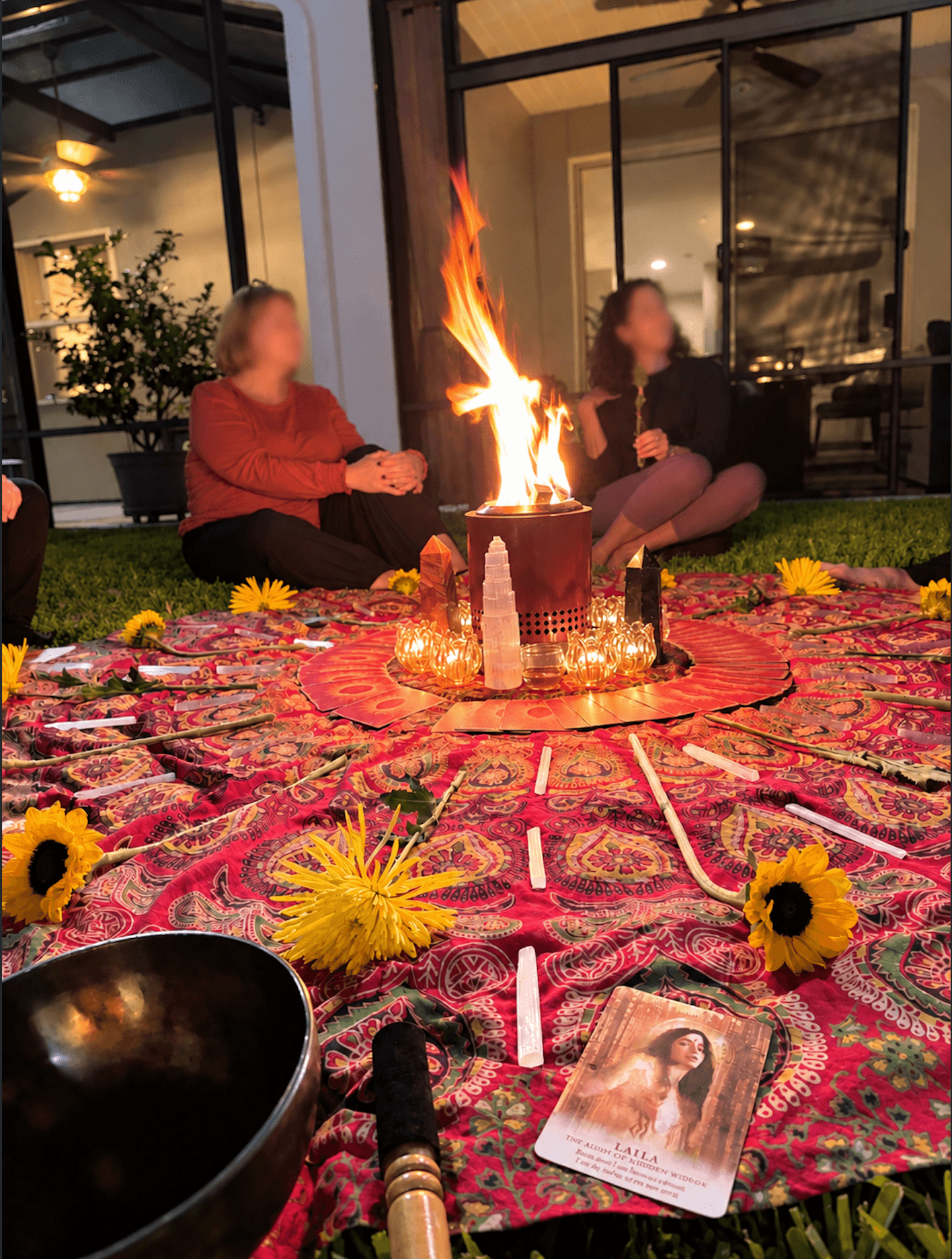 Two women sit on a patio near a fire in a bowl, with a colorful cloth, flowers, and a book on the ground.