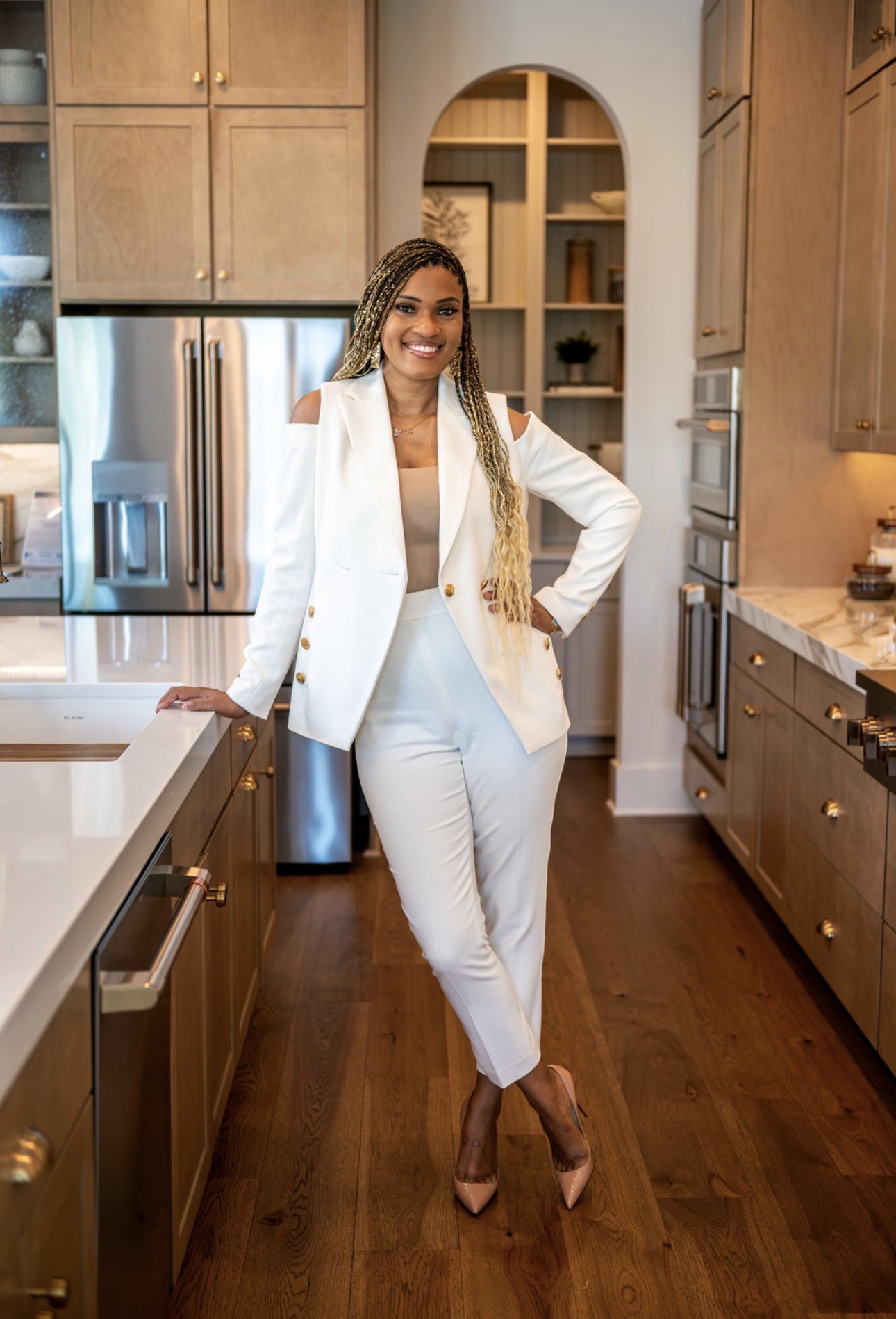 Woman in white suit standing in a kitchen, smiling, with wooden cabinets and stainless steel appliances behind her.