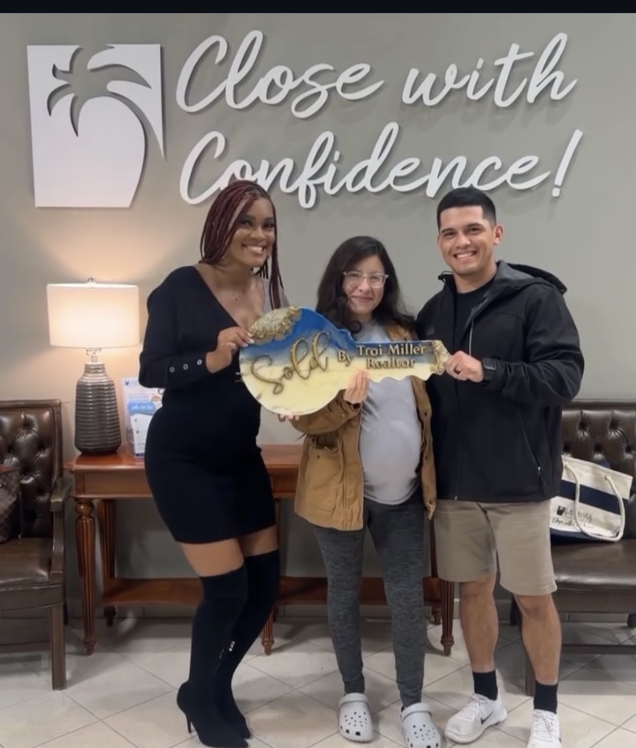 Three people standing indoors, holding a large key, smiling, with a sign that says 'Close with Confidence!' behind them.