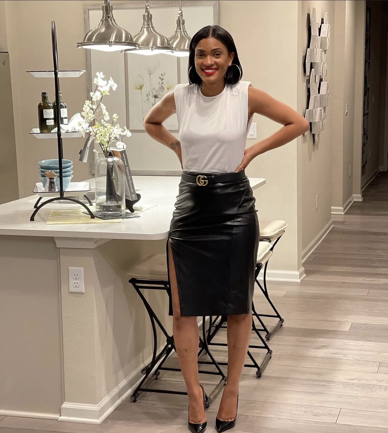 Woman standing in a kitchen, smiling, wearing a white top and black leather skirt, with hands on hips, in a modern home.