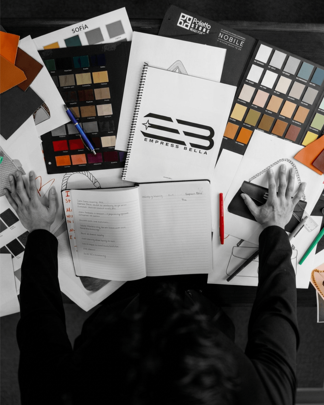 Person working at desk with color swatches, notebooks, and a laptop, viewed from above, in black and white.