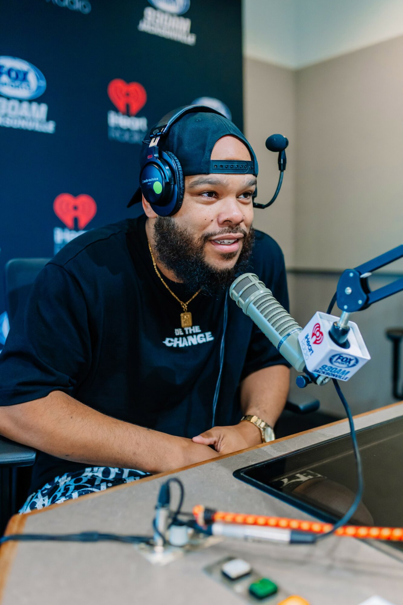 Man wearing headphones and cap speaking into a microphone during a radio interview, smiling, in a studio setting.