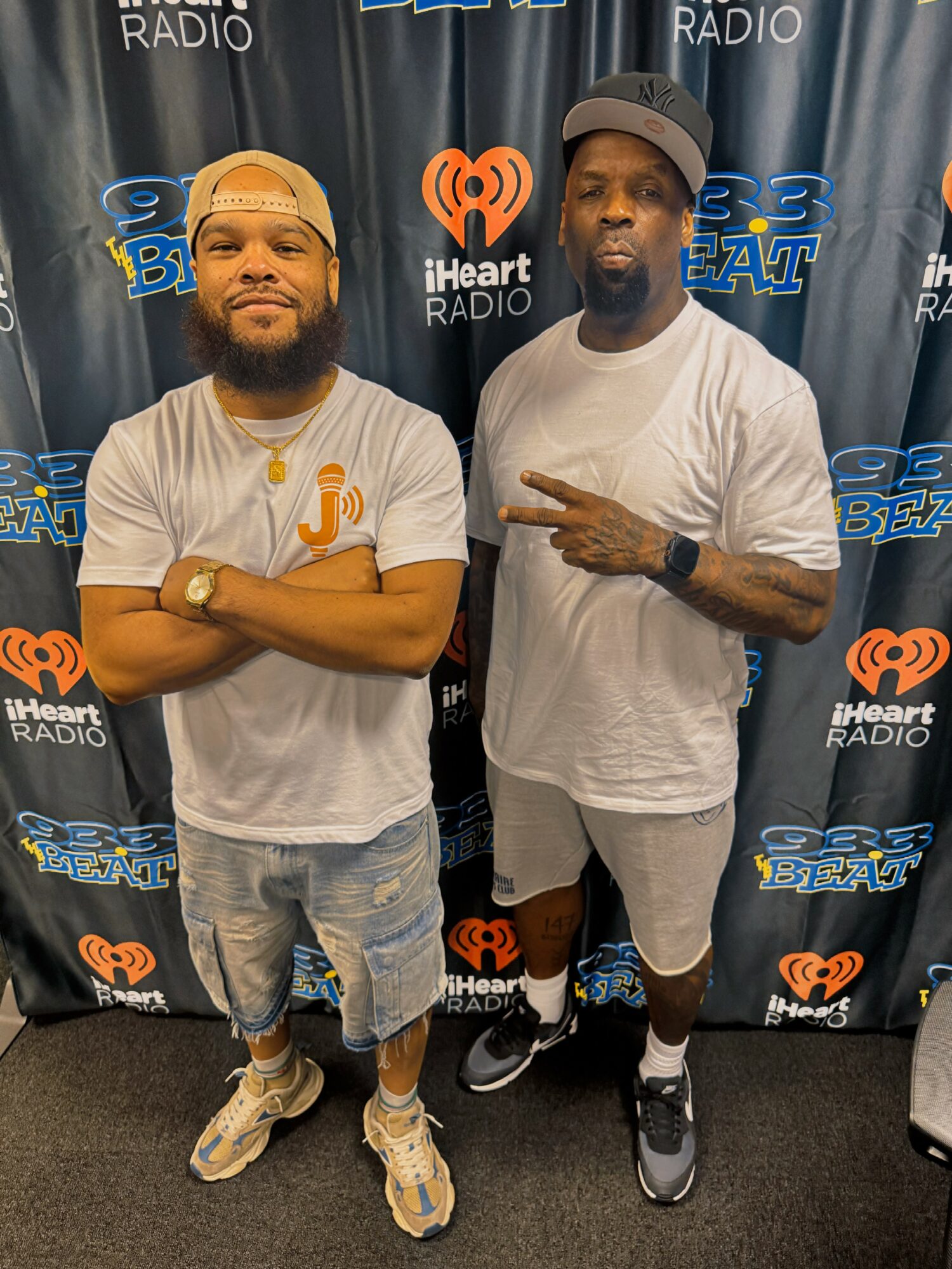 Two men standing in front of a backdrop with radio station logos, one with arms crossed, the other making a peace sign.