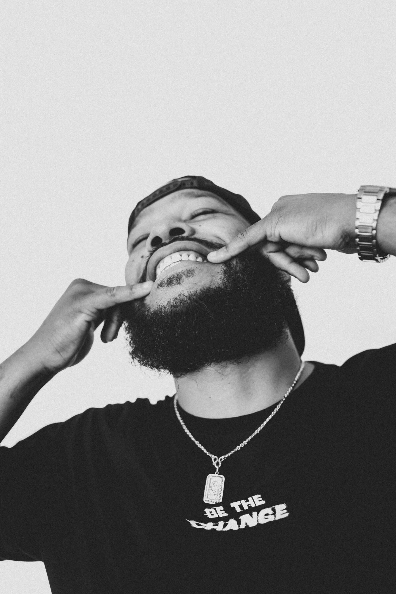 Person with a beard smiling, fingers pulling cheeks, wearing a watch and necklace, black shirt with text, plain background.